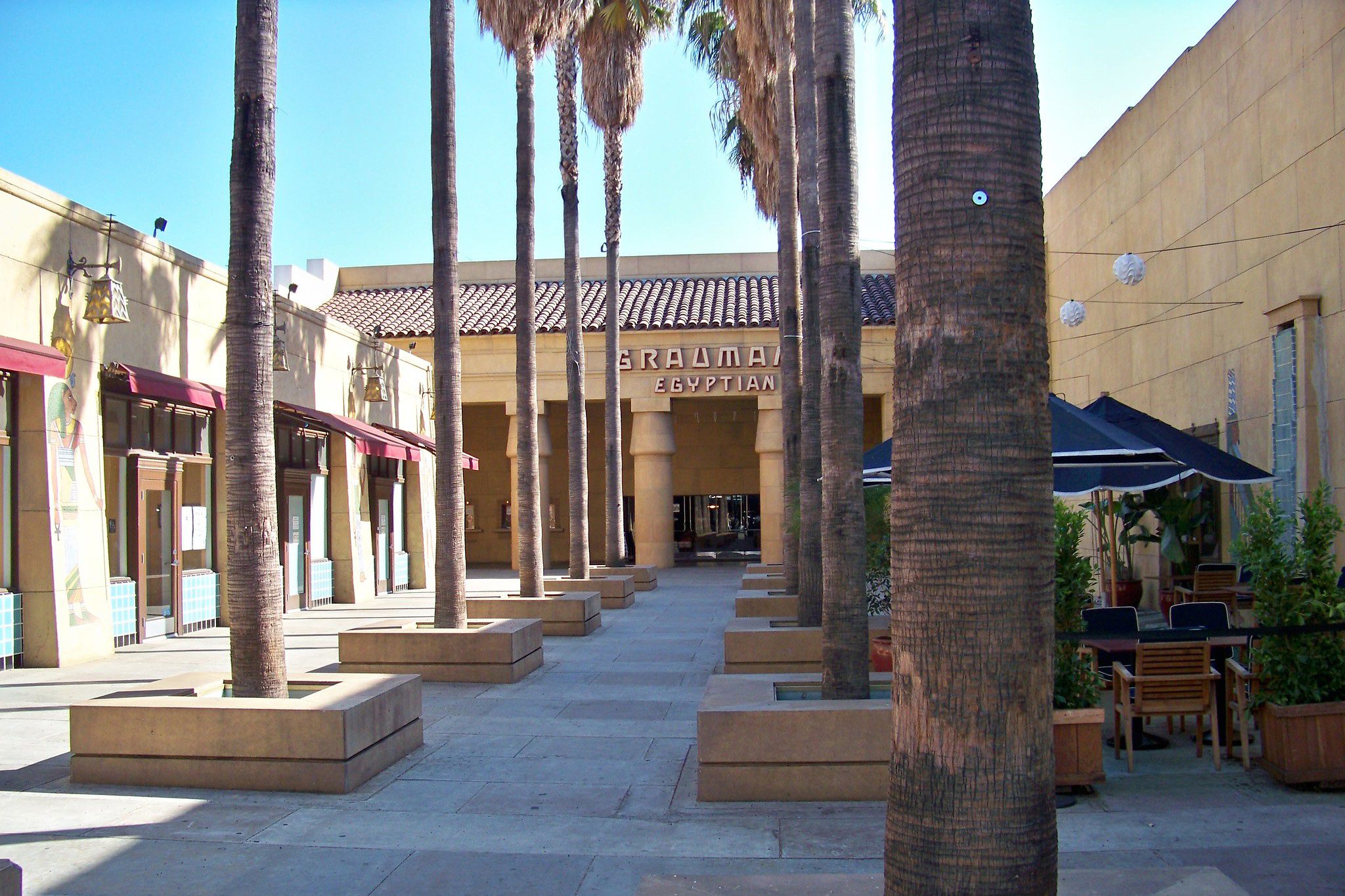 Palm trees line a courtyard leading to the entrance of the Egyptian Theatre, which has tan walls, large columns, and a sign reading "Grauman's Egyptian" above the doorway. Tables and chairs sit to the right.
