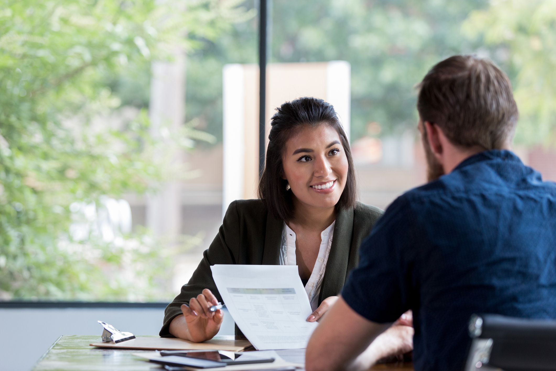 Bank woman smiles while showing a document to a male customer, backside of customer in view, both are sitting at a desk, window showing greenery in the background
