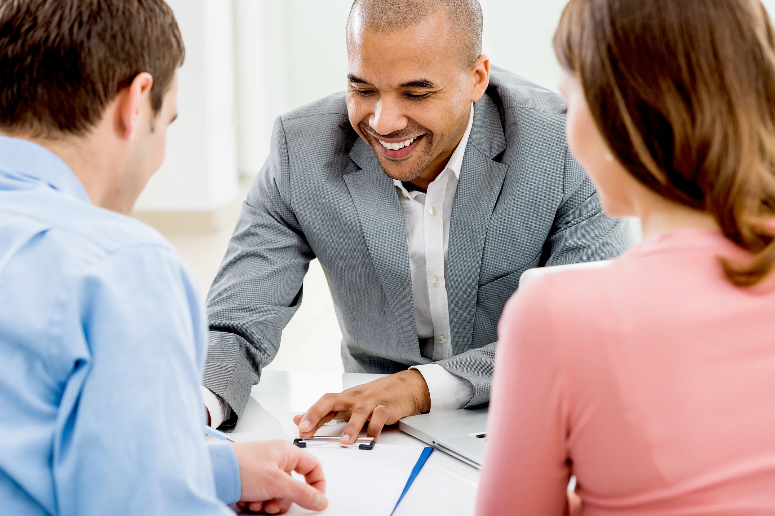Couple meeting with a smiling male mortgage advisor showing paper, at a desk backs of couple showing, mortgage advisor facing front in the middle with his finger pointing to print on paper work