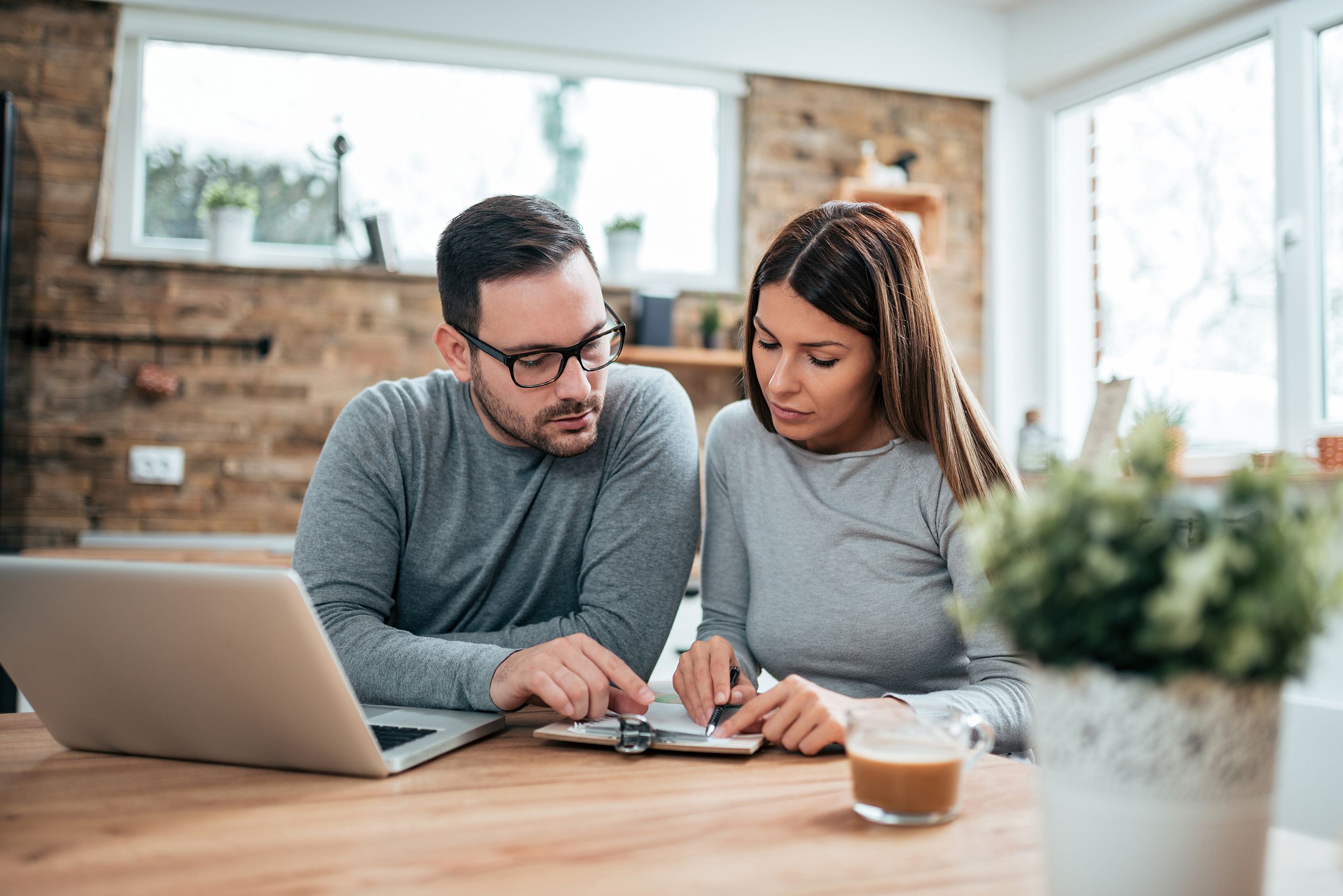 Couple sitting at table doing finances at home with laptop and clipboard, blurred living room in background