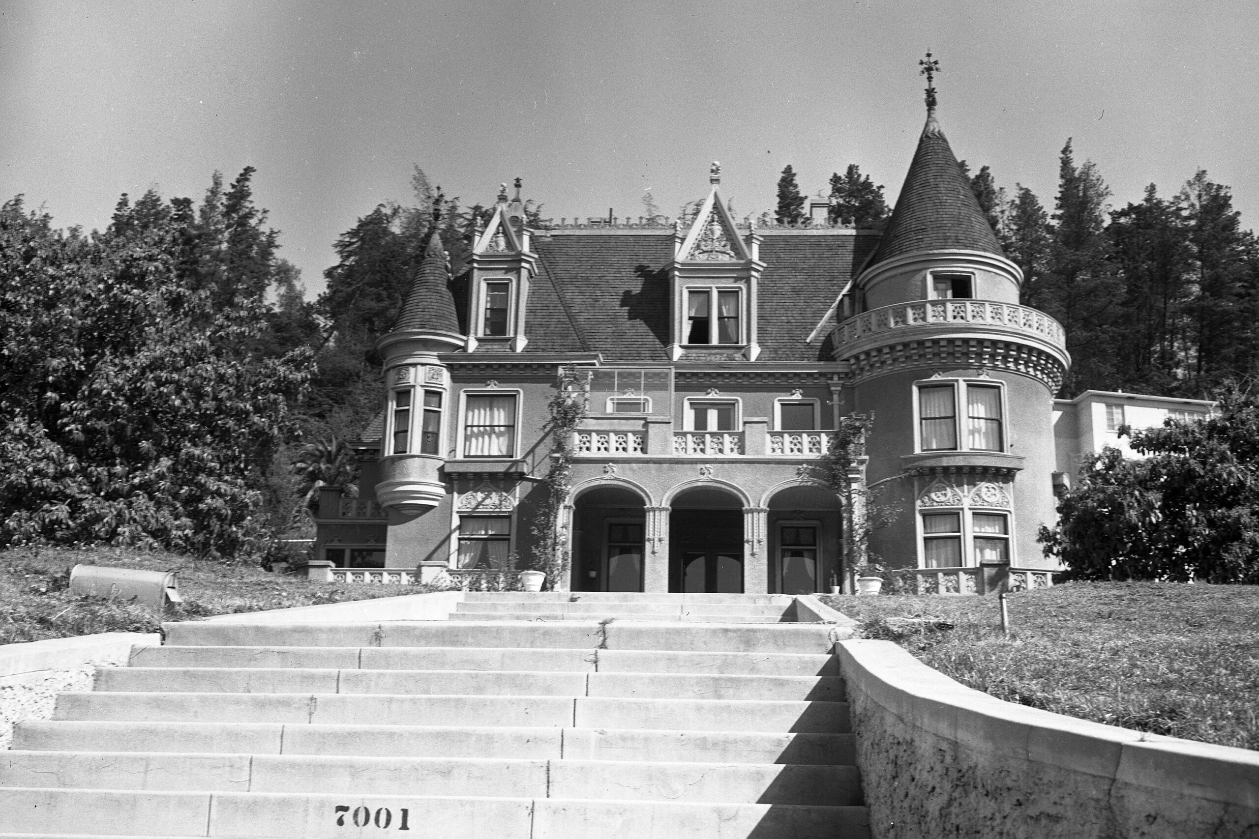 Black and white photo of a large, ornate mansion with turrets, arched windows, and detailed stonework. The house sits at the top of a wide staircase and is surrounded by trees and greenery.