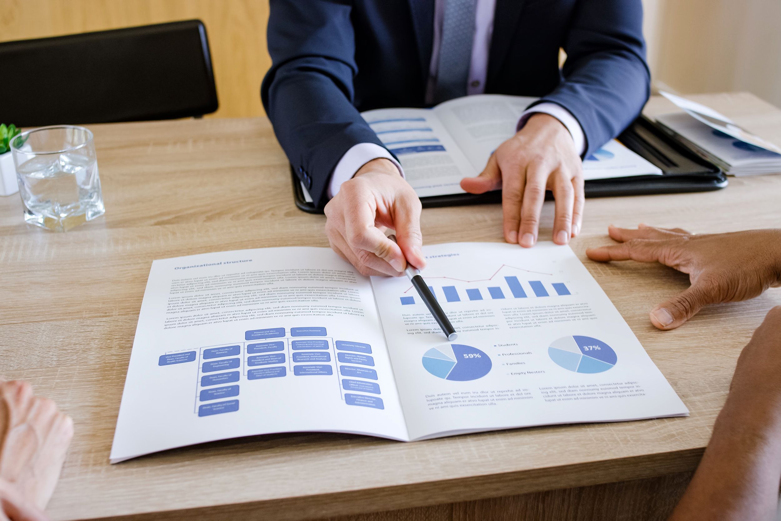 Financial adviser sharing documents with clients, his pen is pointing to pie chart on document on table, focus on hands and arms of financial adviser and clients