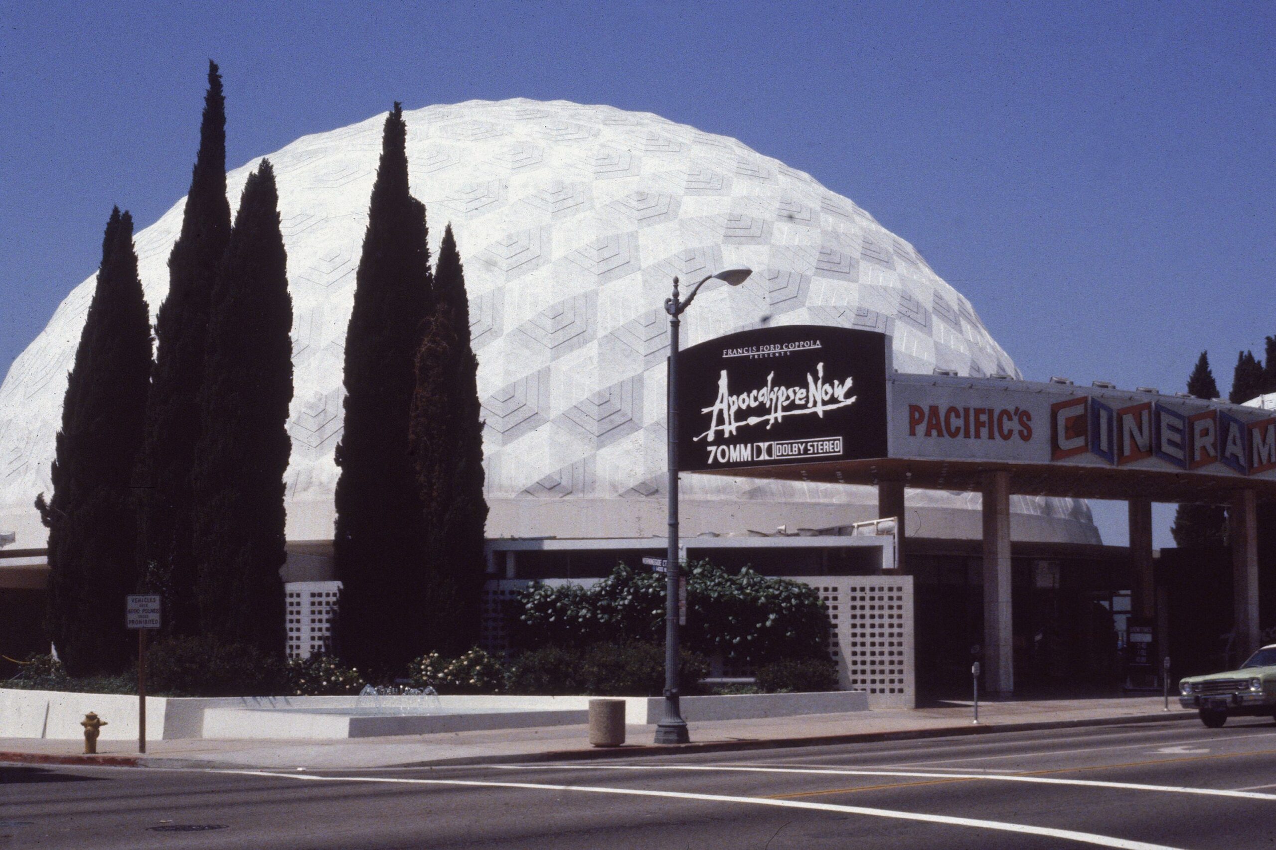 A large white geodesic dome, known as the Cinerama Dome, stands behind trees. A marquee advertises "Apocalypse Now" in 70mm. The sign reads "Pacific’s Cinerama." The scene is taken from across the street.