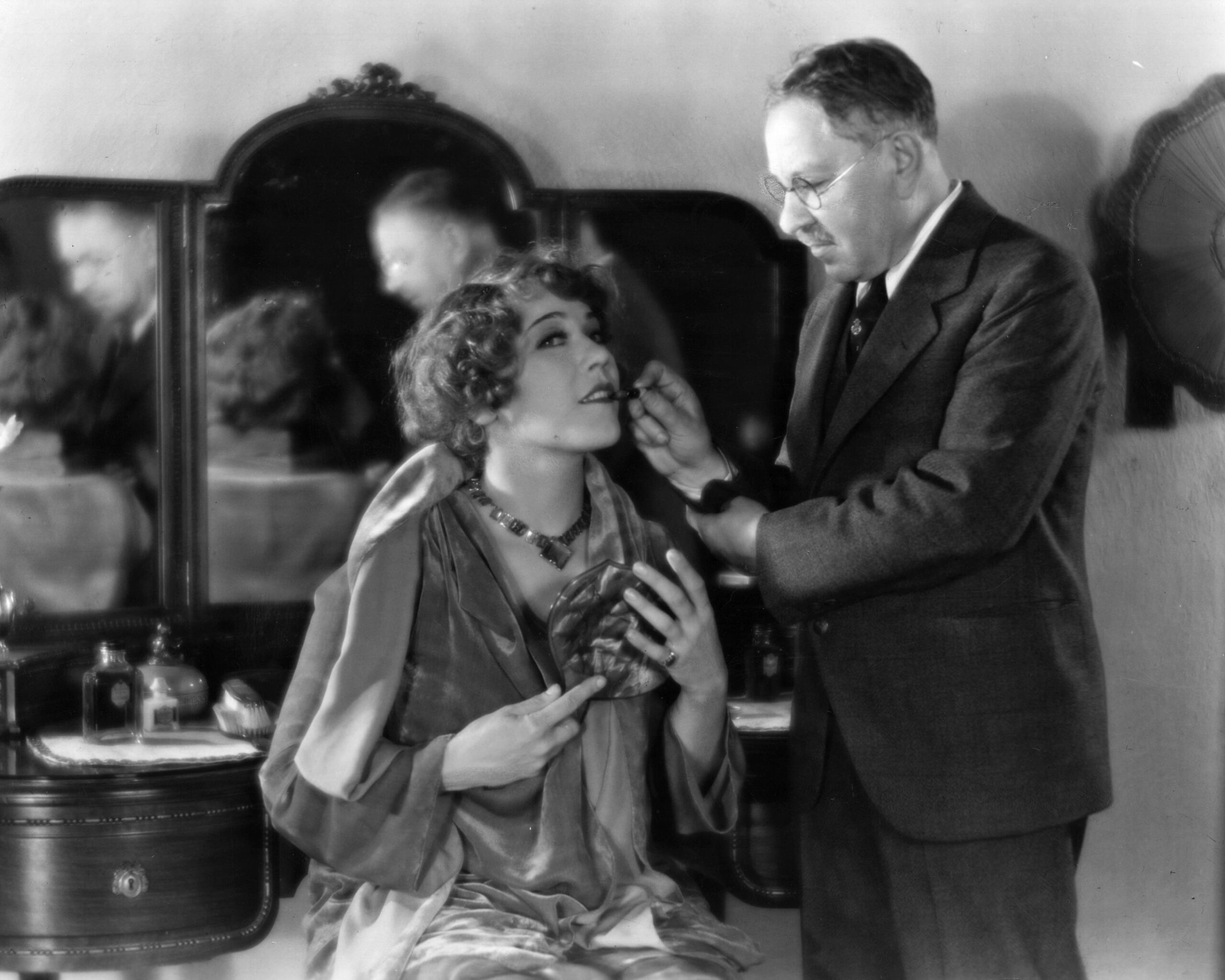 A woman sits at a vanity holding a mirror as a man in a suit applies makeup to her face. Both are focused on the task, with vanity mirrors and bottles visible in the background. The scene appears vintage or early 20th century.