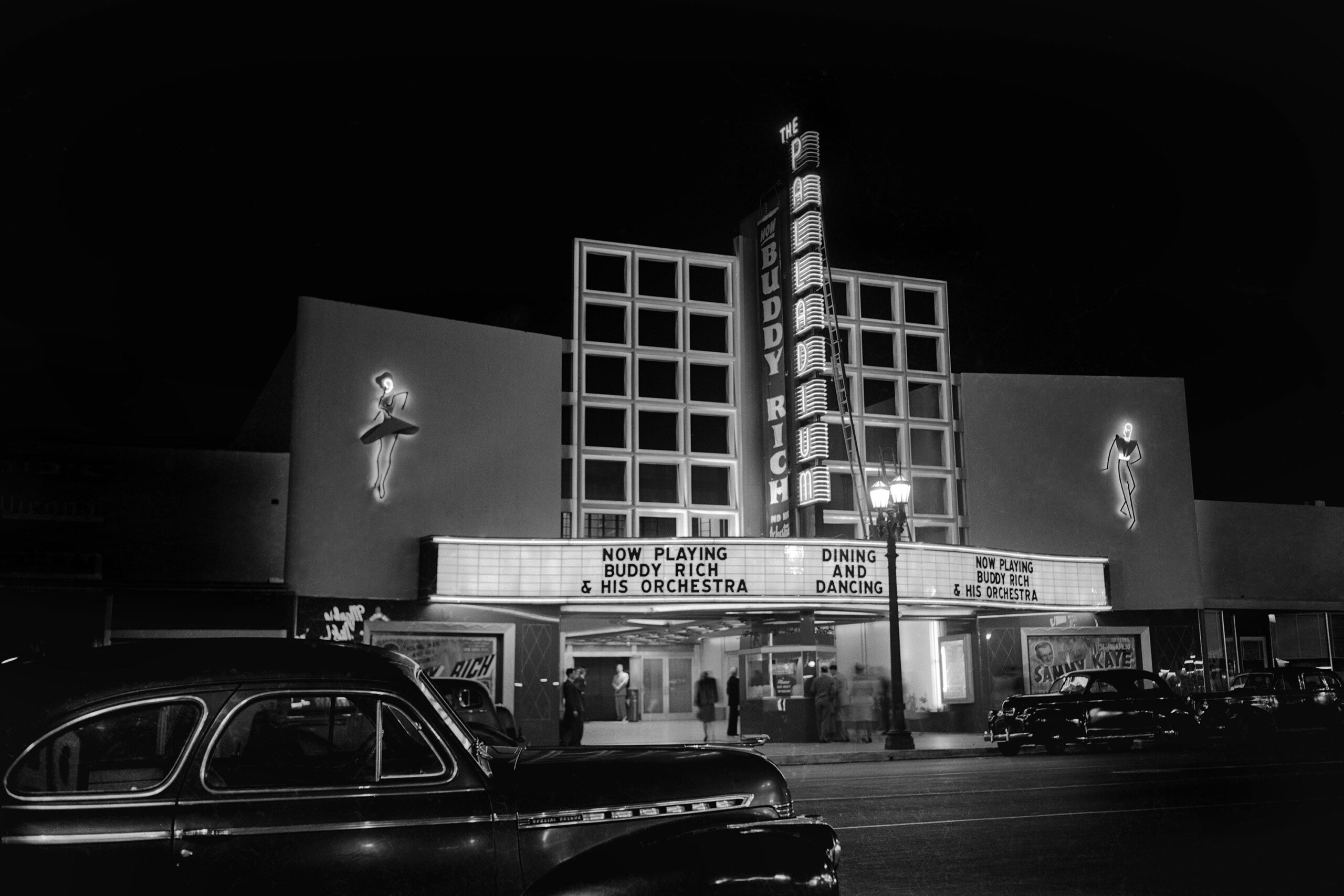 Black-and-white photo of a vintage theater at night, with a brightly lit marquee announcing “NOW PLAYING BUDDY RICH & HIS ORCHESTRA.” Classic cars are parked in front; people stand near the entrance under neon lights.