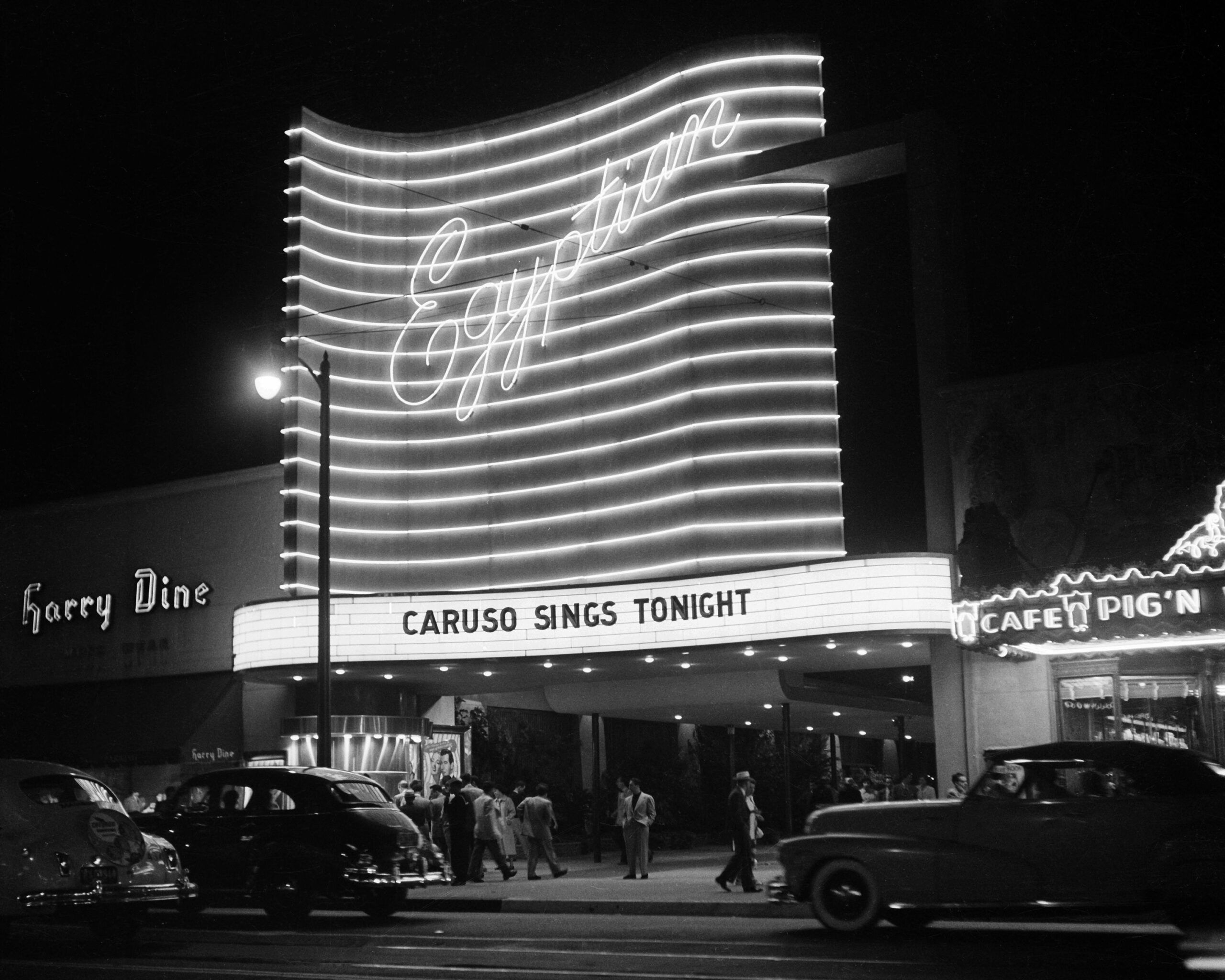 A nighttime street scene shows a crowd outside the illuminated Egyptian Theatre. The marquee reads “CARUSO SINGS TONIGHT.” Classic cars are parked along the curb. Neon lights and nearby shops are visible.