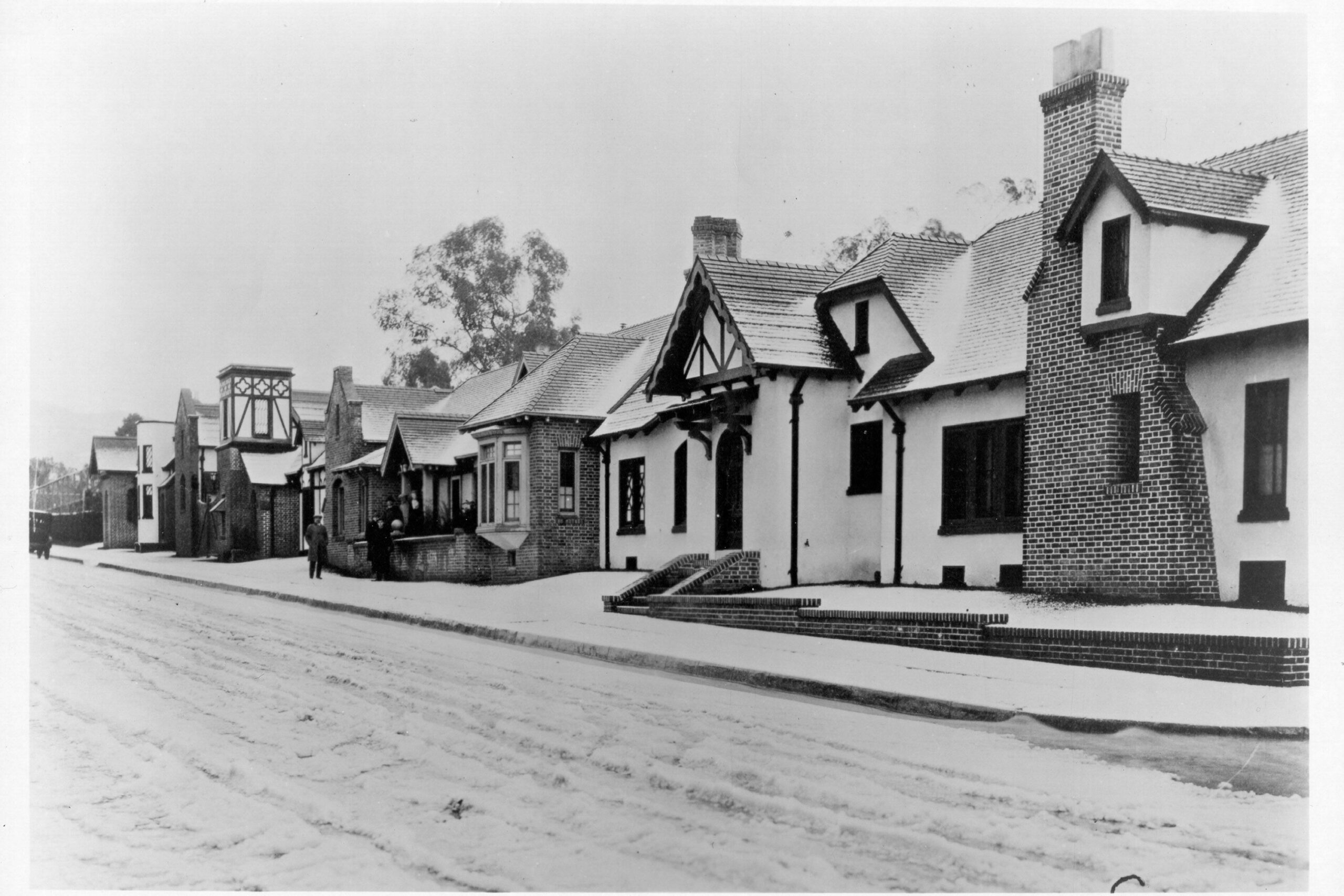 Black and white photo of a quiet residential street lined with houses featuring steep, gabled roofs, chimneys, and brickwork. The sidewalk and road appear empty and the scene looks peaceful.