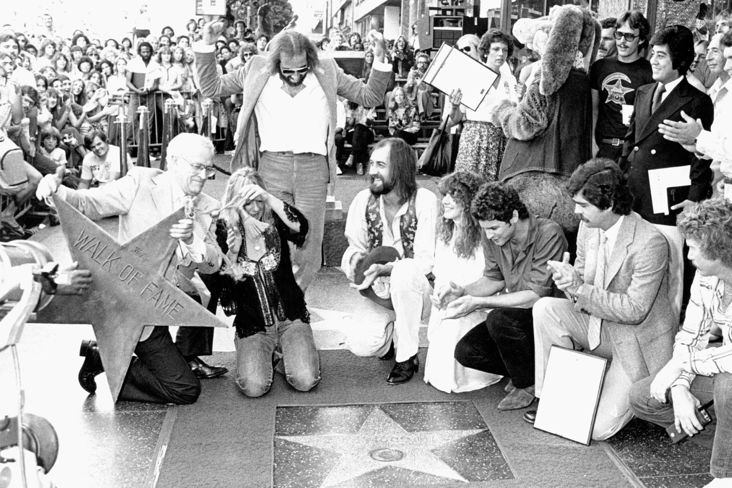 A group of people celebrate at a Hollywood Walk of Fame ceremony, gathered around a star on the sidewalk, with a crowd watching and some holding a large star-shaped plaque.