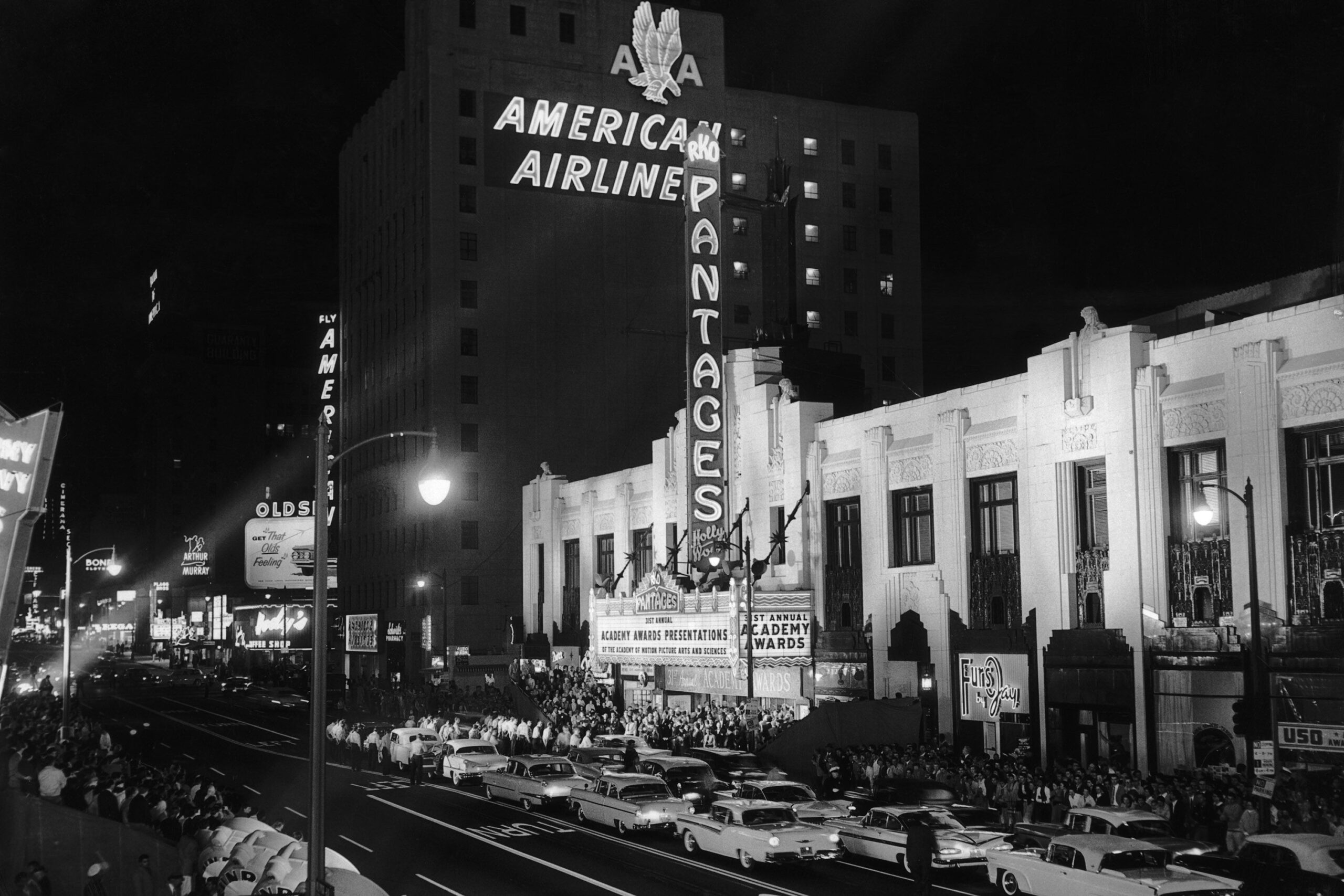 Black-and-white photo of a busy nighttime street outside the Pantages Theatre, with crowds and vintage cars; a large sign reads “ACADEMY AWARDS” and neon lights illuminate “AMERICAN AIRLINES” and “PANTAGES.”
