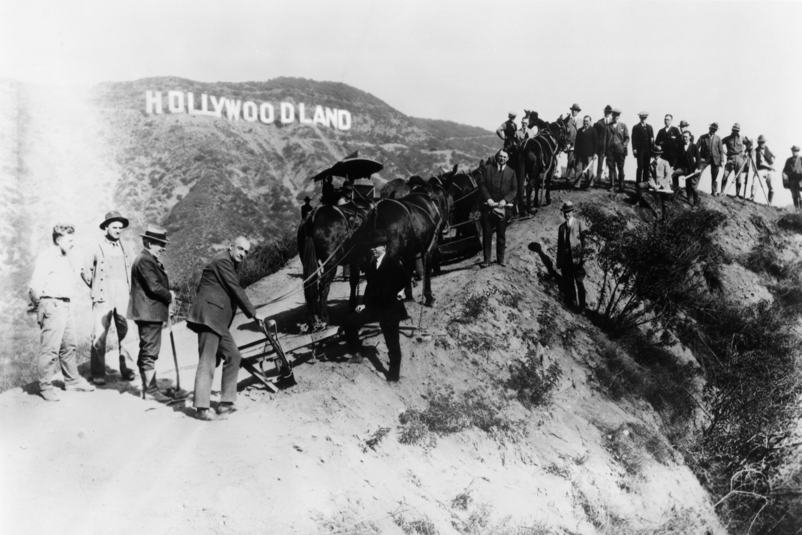 Black and white photo of people and horses on a hillside near the original "Hollywoodland" sign, with several men standing or sitting, and a group of riders on horseback in the background.