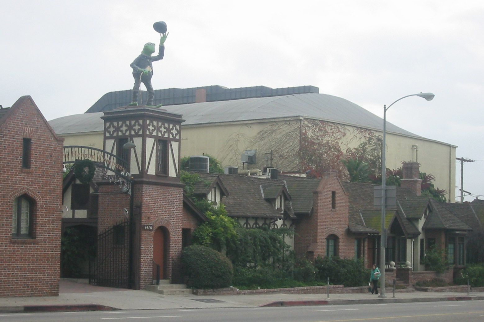 A whimsical building with Tudor-style architecture featuring a statue of Kermit the Frog tipping his hat, perched above an arched entrance. Trees and a person walking are visible nearby.