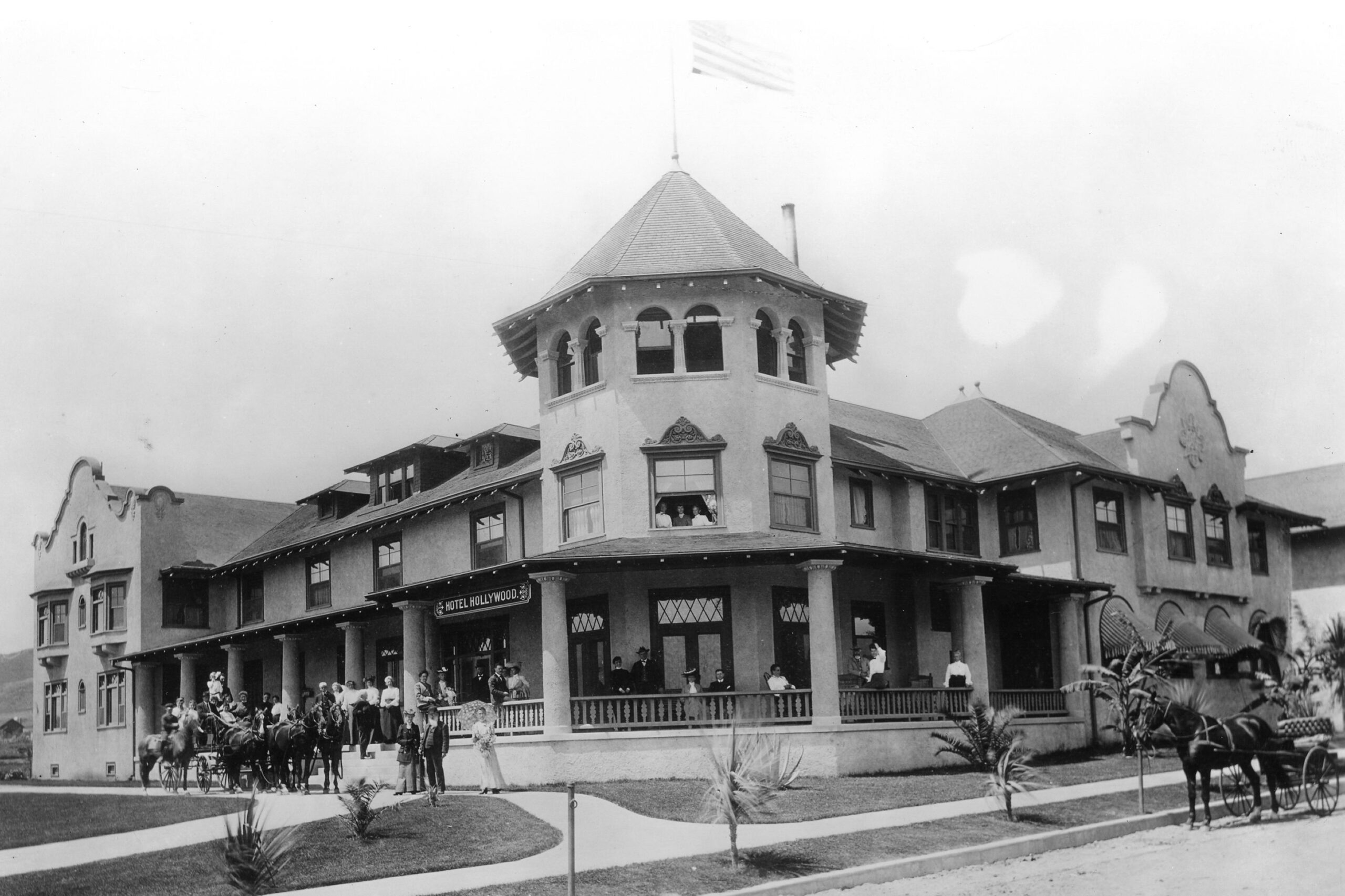 Historic black-and-white photo of a large, two-story hotel with a central turret and an American flag on top. People and horse-drawn carriages are gathered outside the building.