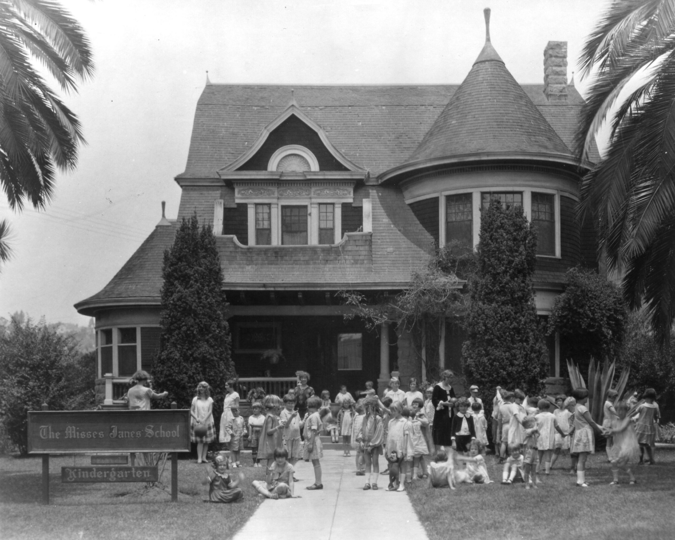 A group of children and adults gather outside a large Victorian-style house with a turret and front porch. A sign reads "The Misses Janes School - Kindergarten." Palm trees frame the scene.