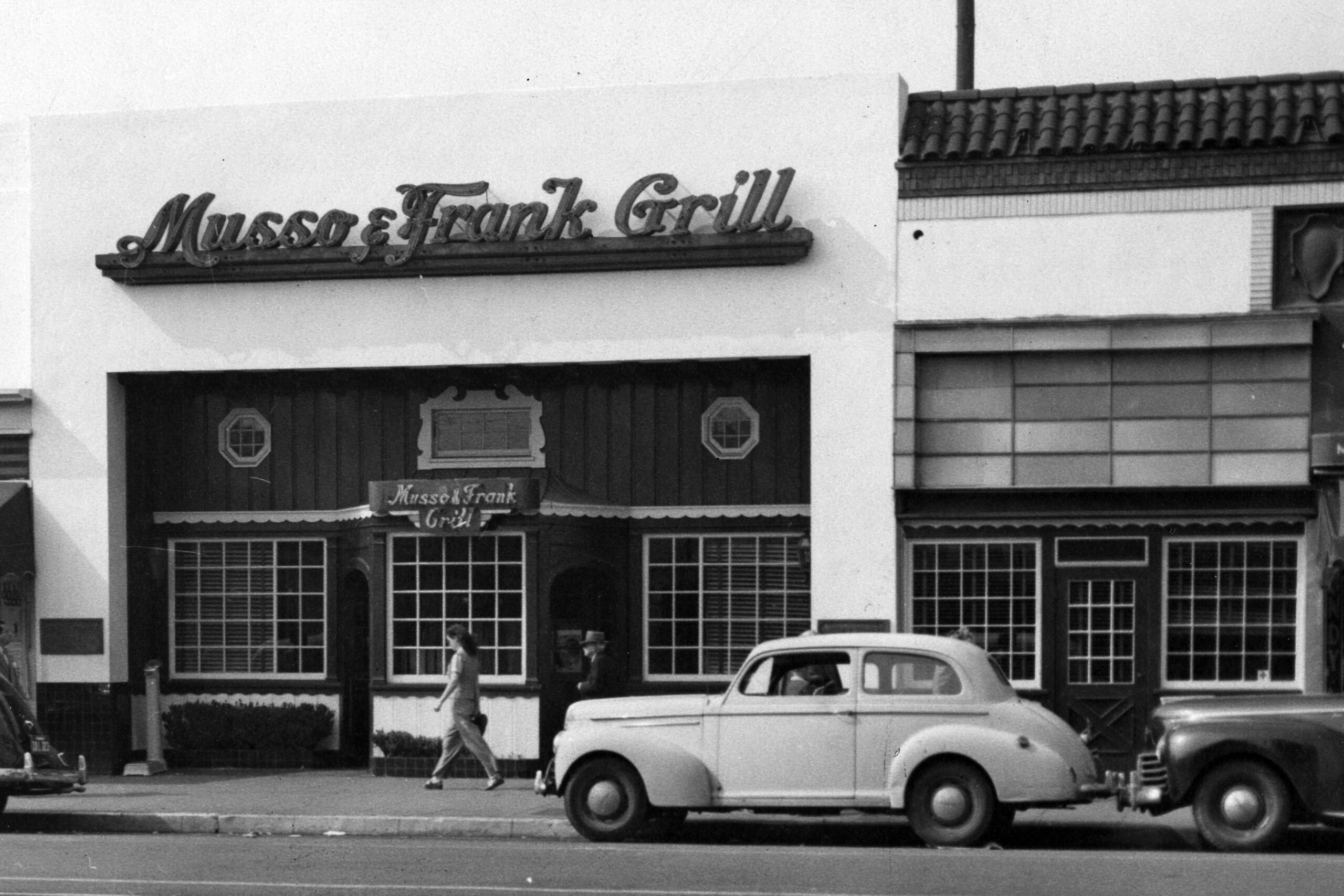 Black and white photo of the Musso & Frank Grill restaurant exterior, featuring its sign above the entrance and a vintage car parked in front. A person walks past the building on the sidewalk.
