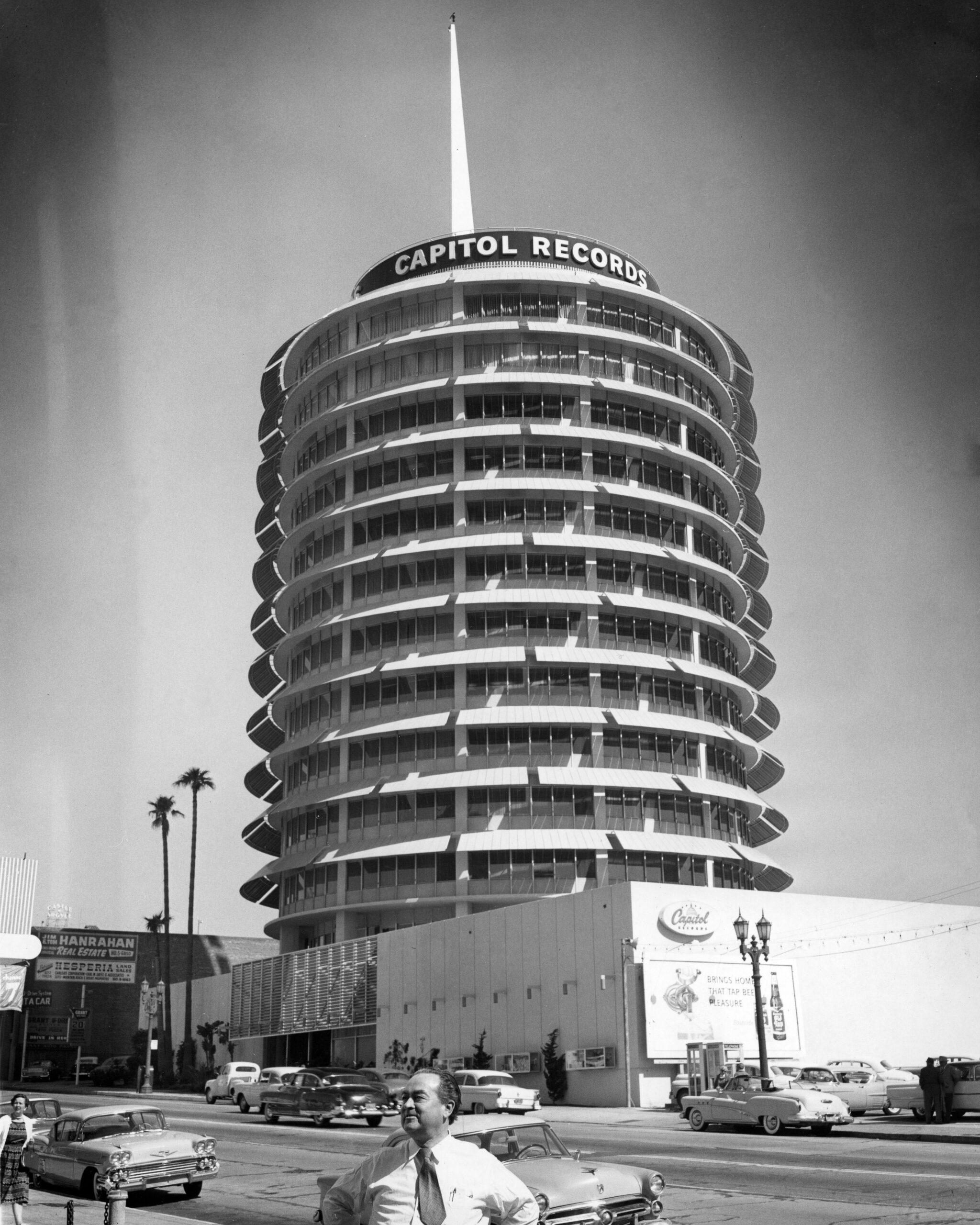 A black-and-white photo of the Capitol Records building in Los Angeles, shaped like a stack of records, with vintage cars parked outside and a man smiling in the foreground.
