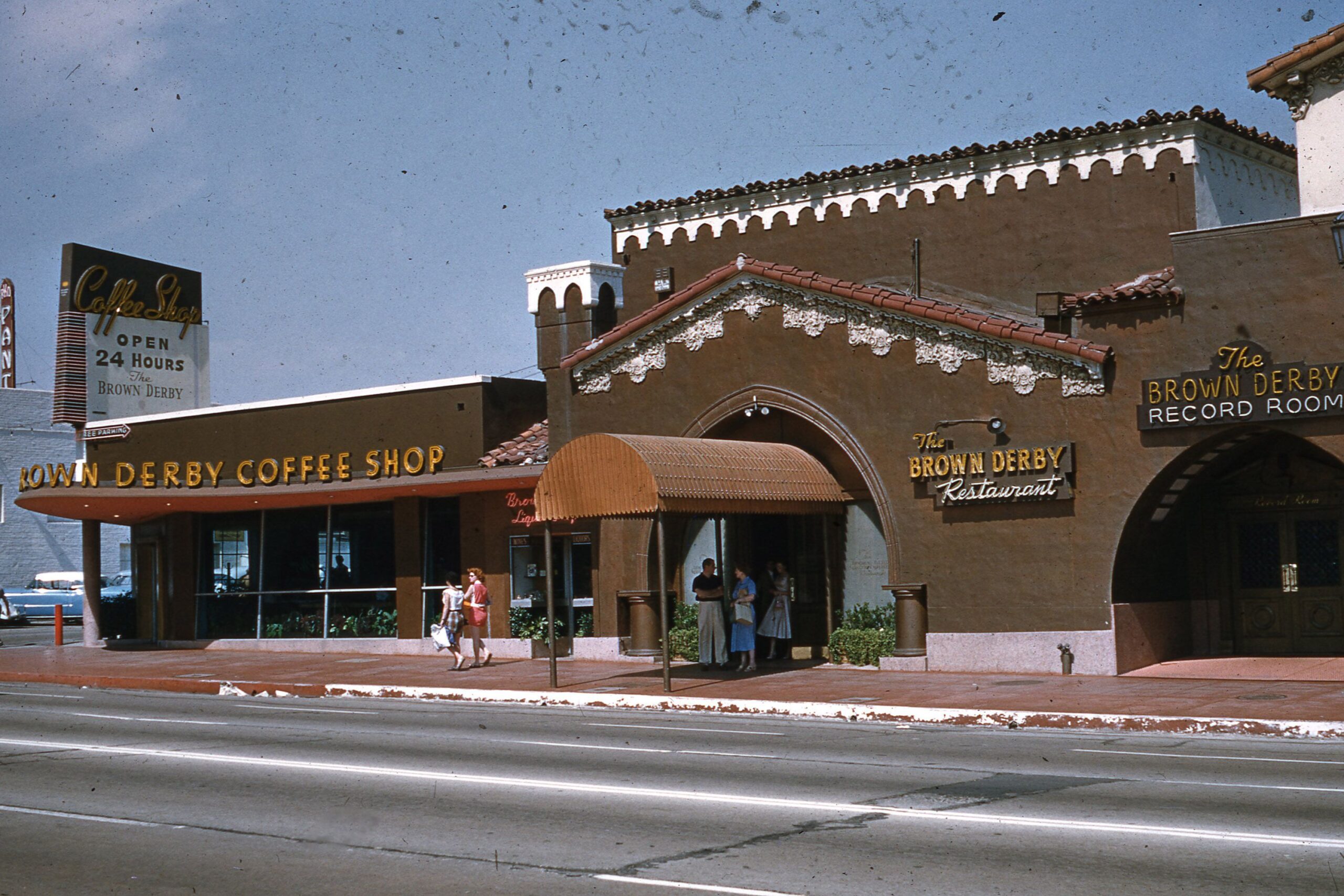 A vintage street view of The Brown Derby Coffee Shop and Restaurant, featuring people entering the building. Signs advertise 24-hour service and a record room. The building has a Spanish-style facade with decorative trim.
