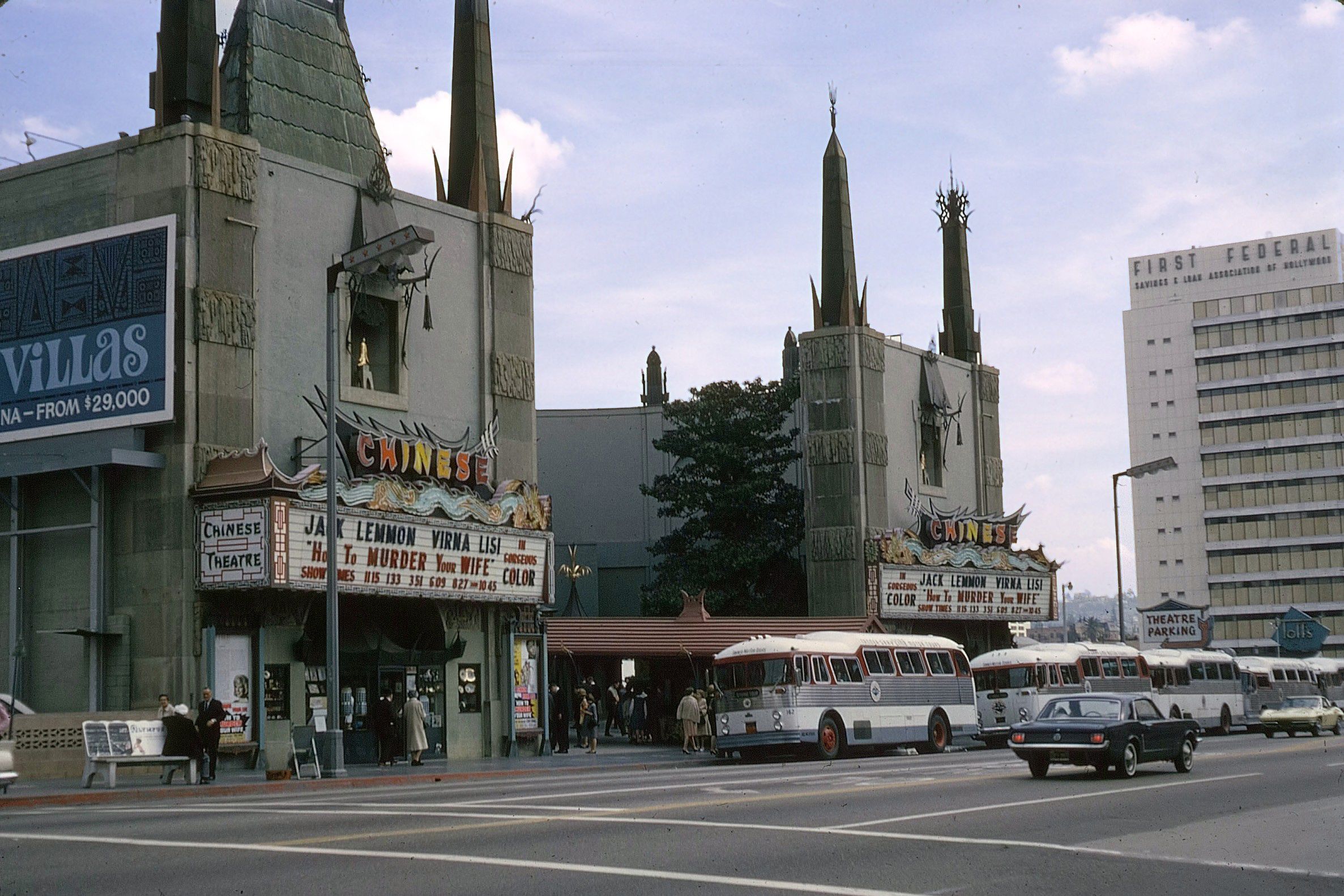 A street view of Grauman’s Chinese Theatre in Hollywood, with vintage buses and cars parked in front, people walking on the sidewalk, and a tall office building in the background. The theater marquee advertises live shows.