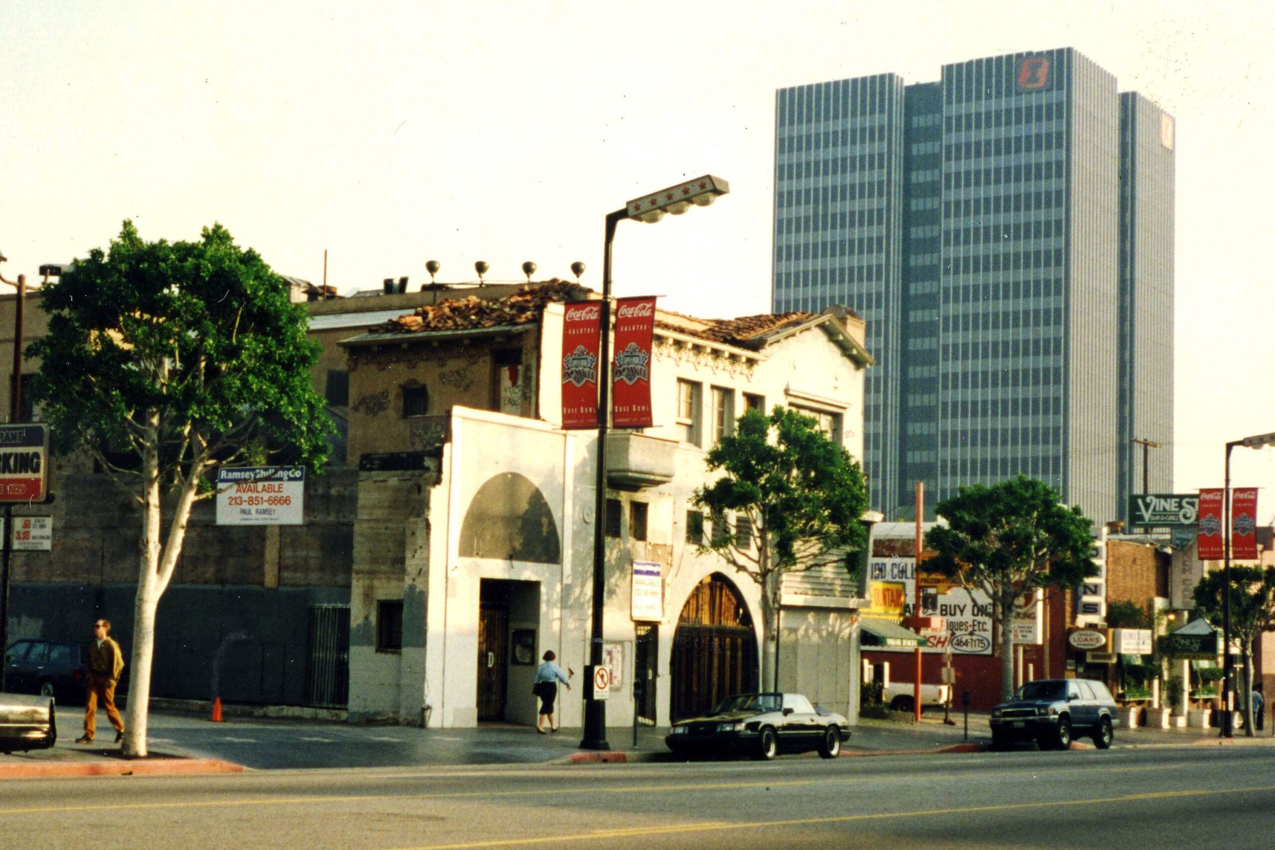 A city street with older, worn buildings in the foreground, modern high-rise offices in the background, and a few cars and pedestrians. Trees line the street and red Coca-Cola banners hang from lampposts.