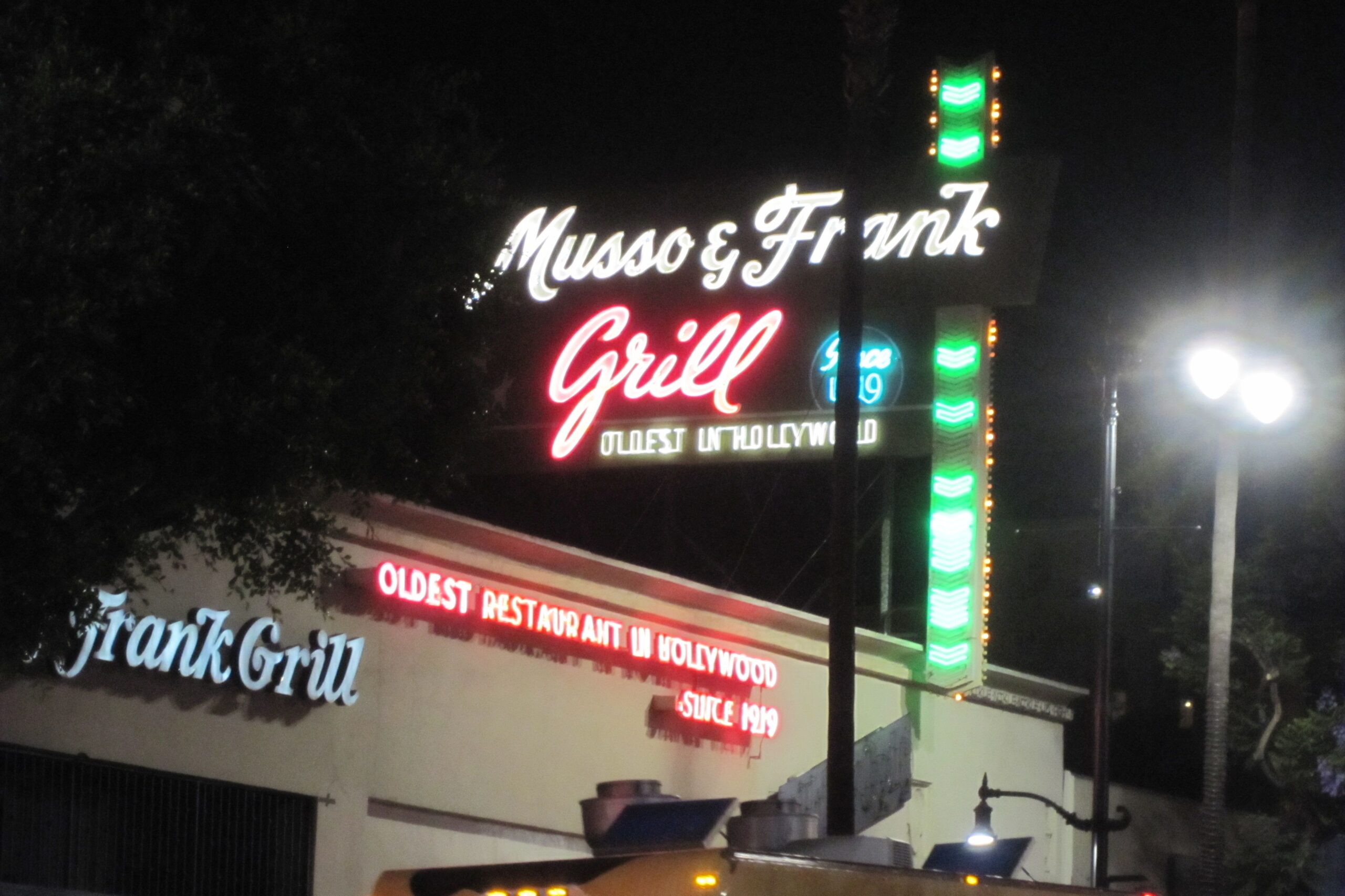 A nighttime view of the Musso & Frank Grill, featuring neon signs advertising it as the oldest restaurant in Hollywood, established in 1919. The building is partially obscured by trees and streetlights.