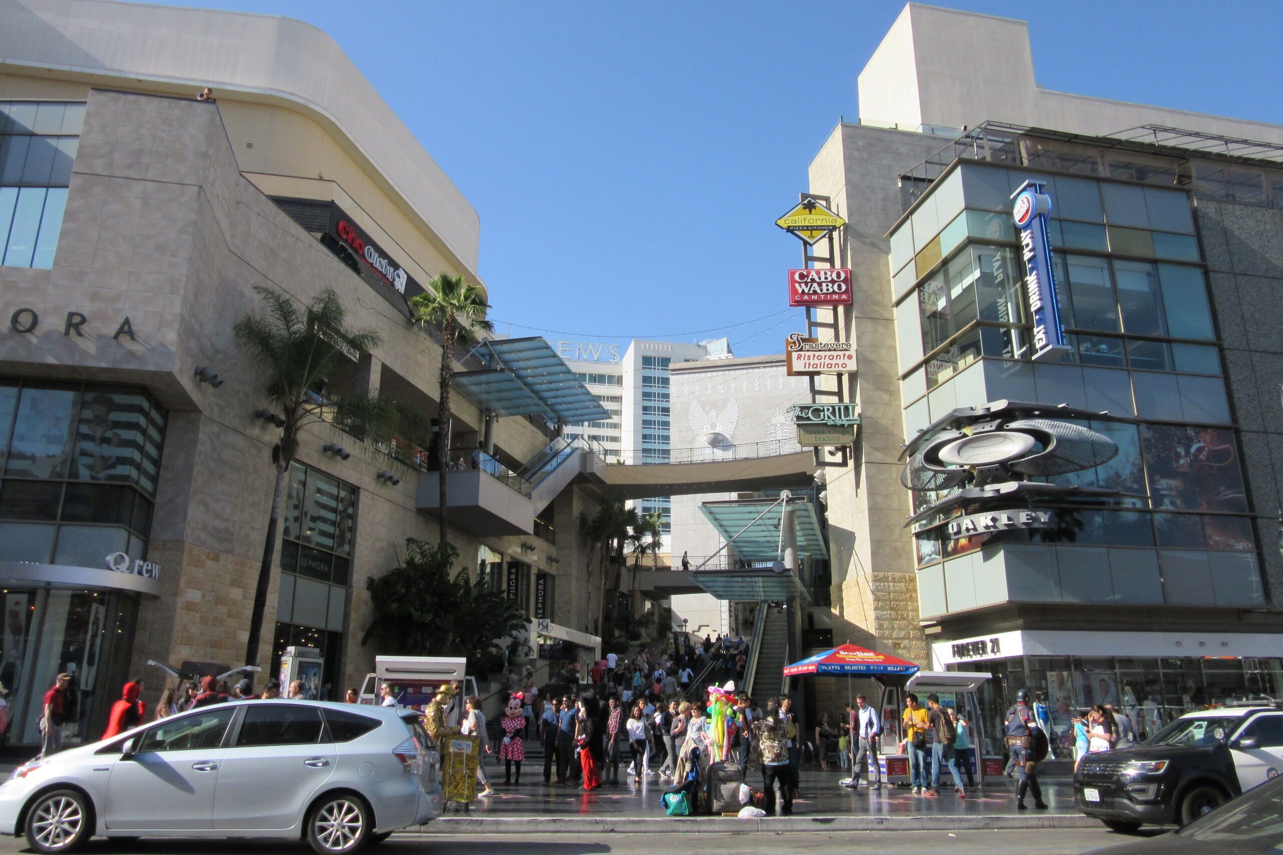 Crowds of people walk outside a busy shopping mall with stores like Sephora, Cabo Wabo, and Oakley. Cars drive past on the street in front, and the sky is clear and blue above the modern buildings.