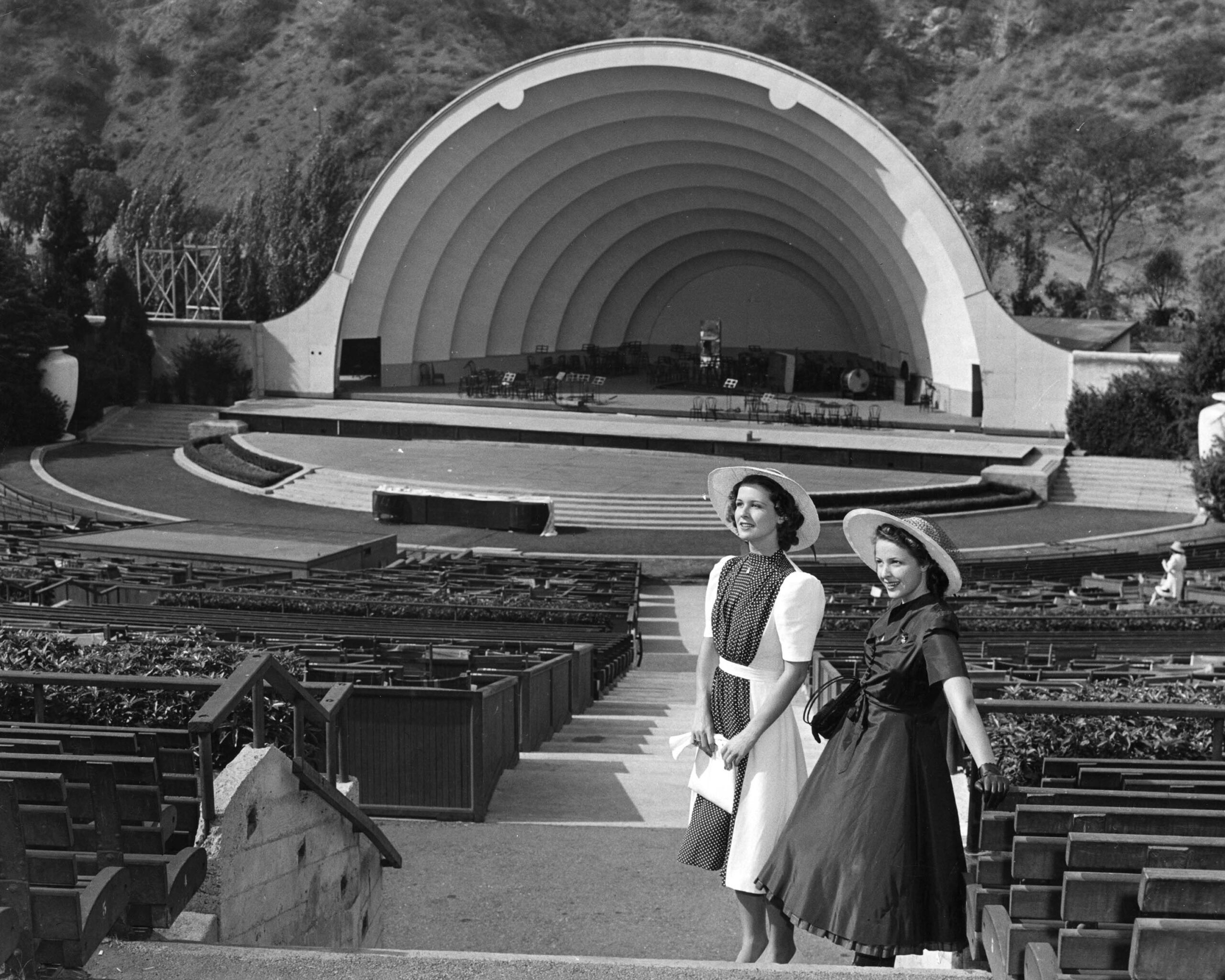 Two women in vintage dresses and hats stand in front of an empty outdoor amphitheater with a large curving shell stage, surrounded by rows of empty seats and trees in the background.