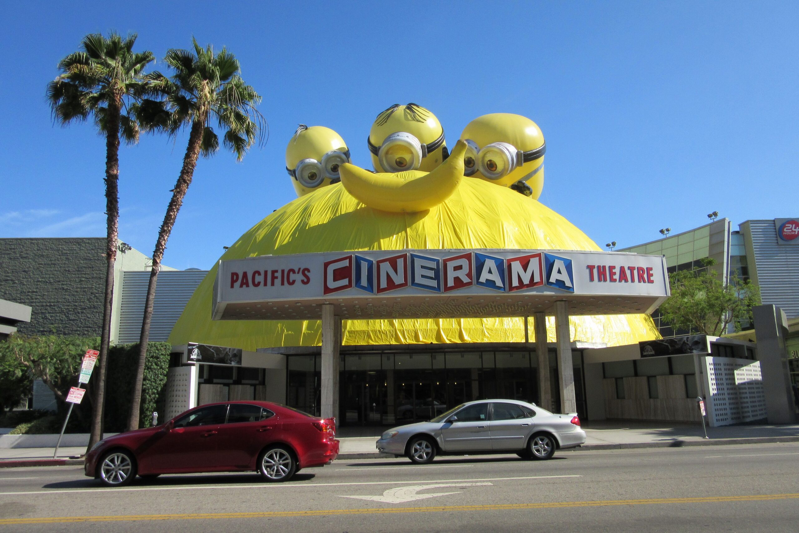 The Pacific’s Cinerama Theatre features a large, dome-shaped structure covered with a giant Minions decoration. Three Minion faces peek over the yellow dome. Two cars are parked on the street in front.