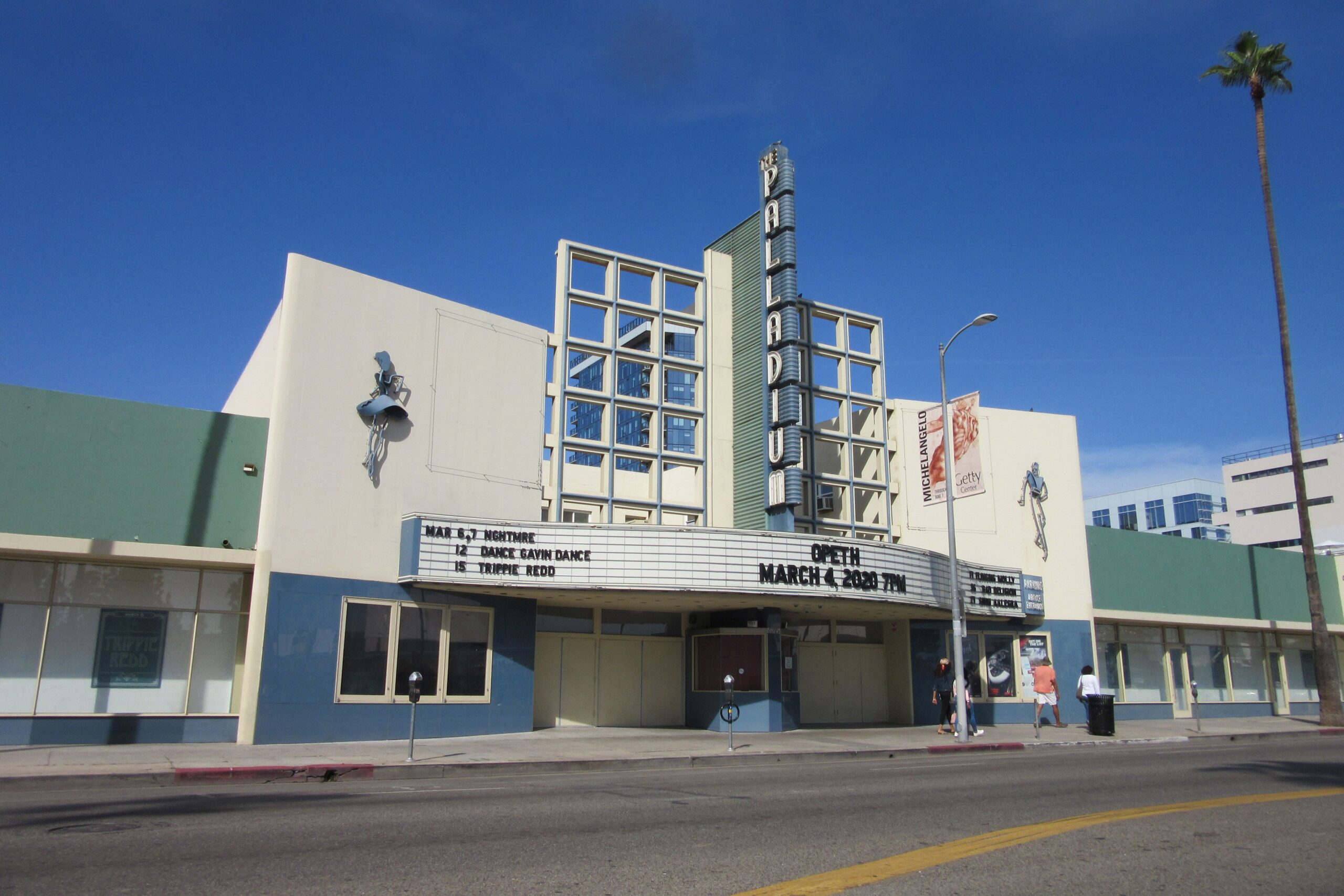 Art Deco-style theater building with a vertical "Paradise" sign, marquee advertising upcoming events, and a few people walking by on a clear, sunny day. A tall palm tree stands to the right of the theater.