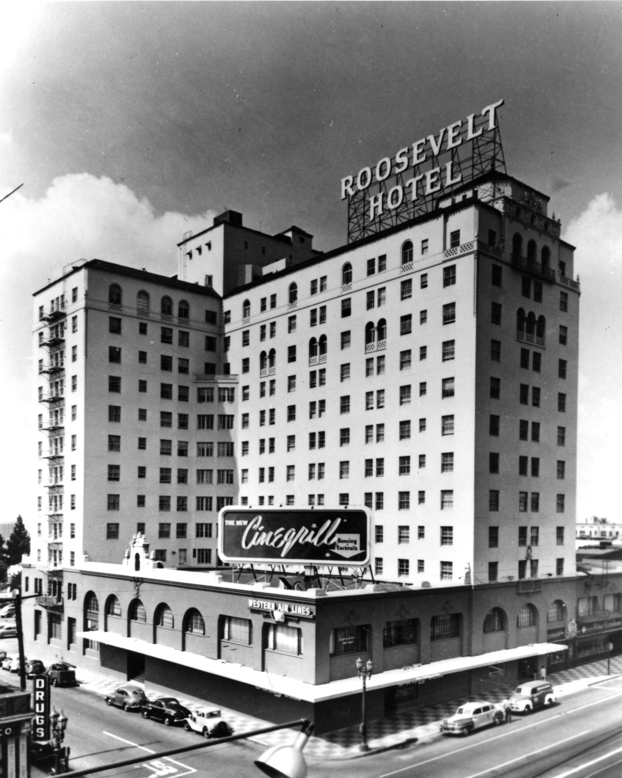 Black-and-white photo of the Roosevelt Hotel, a large multi-story building with prominent rooftop signage, vintage cars parked along the street, and a large “Cinegrill” sign above the entrance.