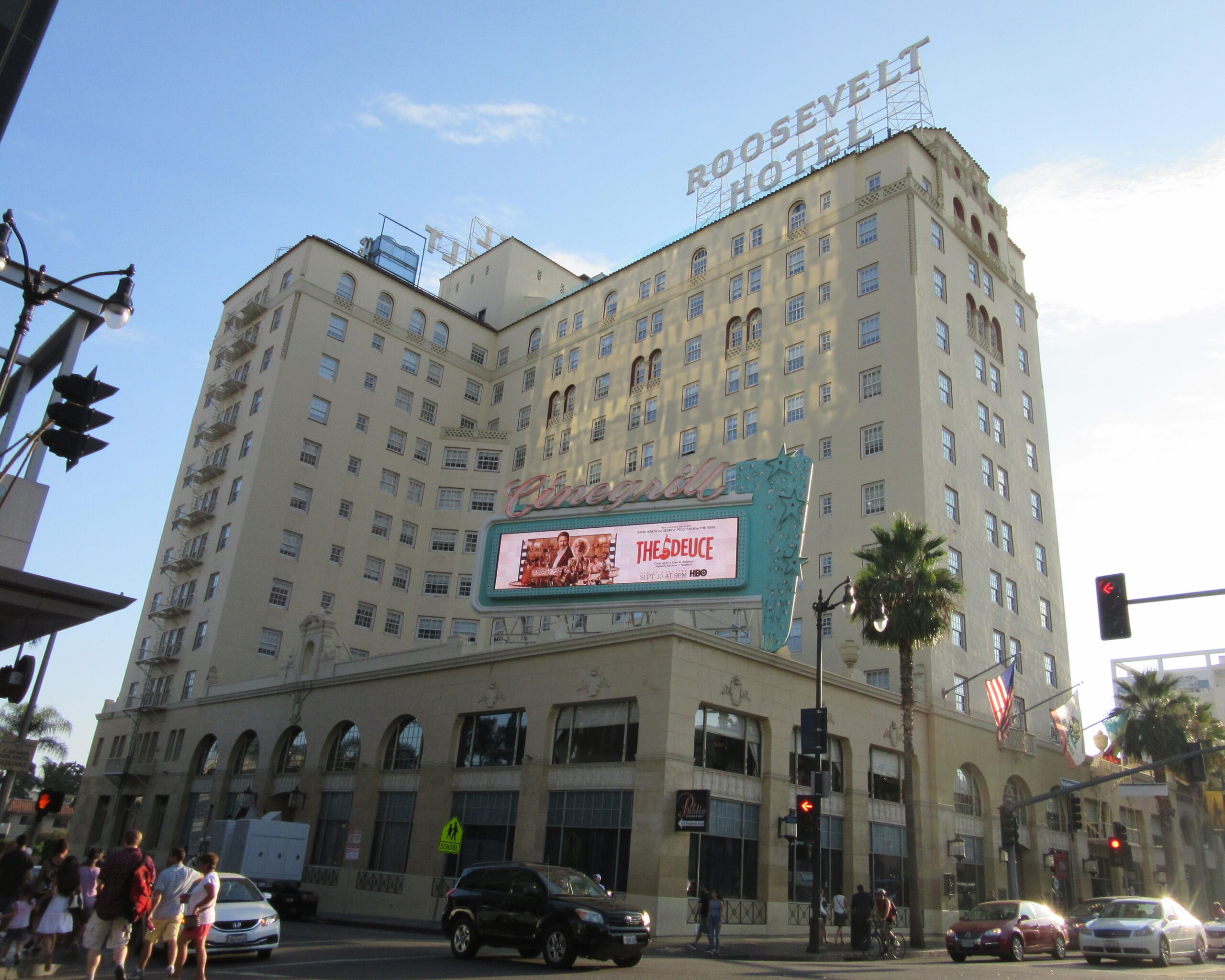 The Hollywood Roosevelt Hotel with a neon sign and digital billboard on the facade, palm trees nearby, American flags, and people crossing the street at a busy Los Angeles intersection.