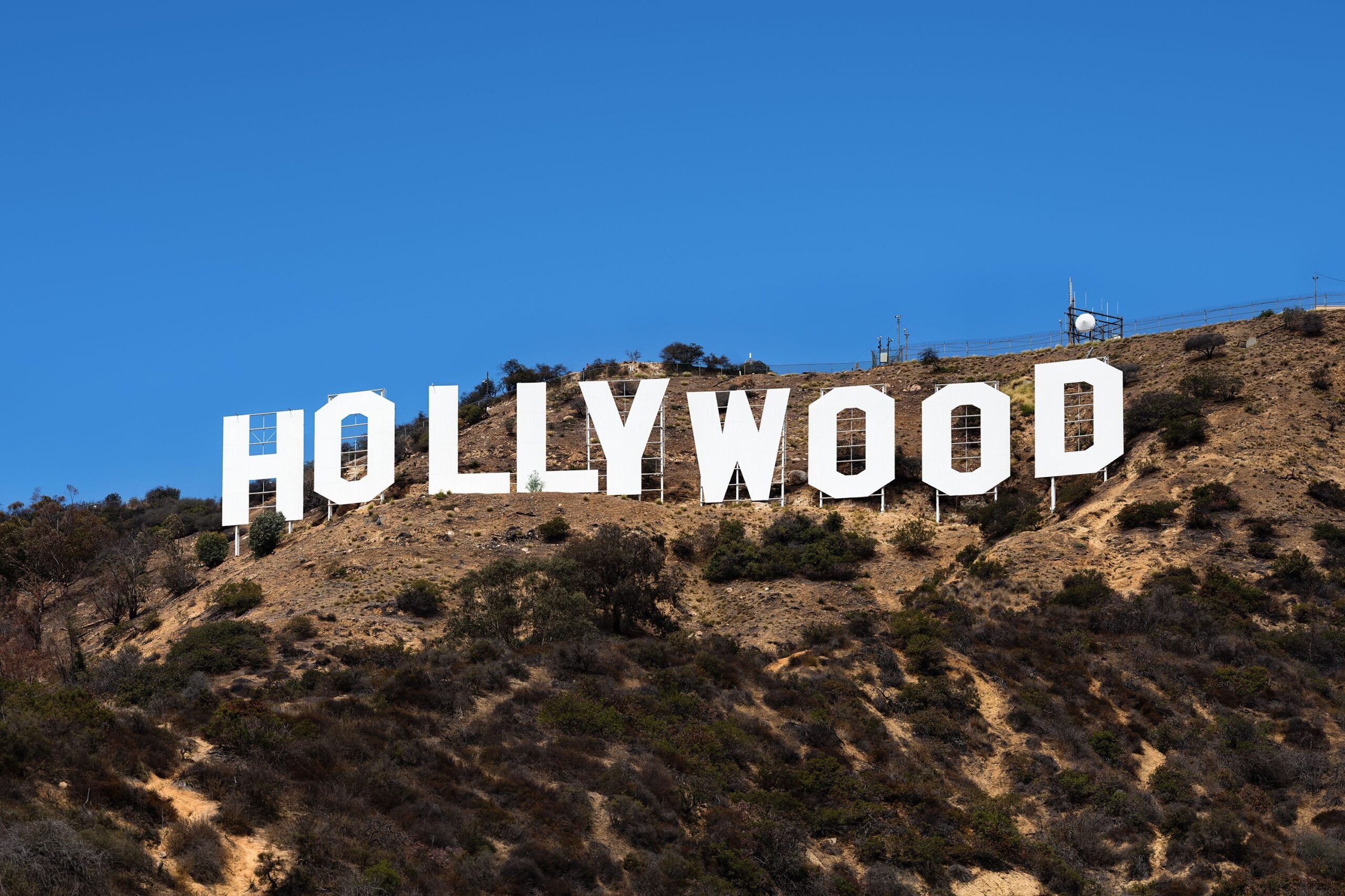 The iconic white Hollywood sign stands on a dry, shrub-covered hillside under a clear blue sky in Los Angeles, California.