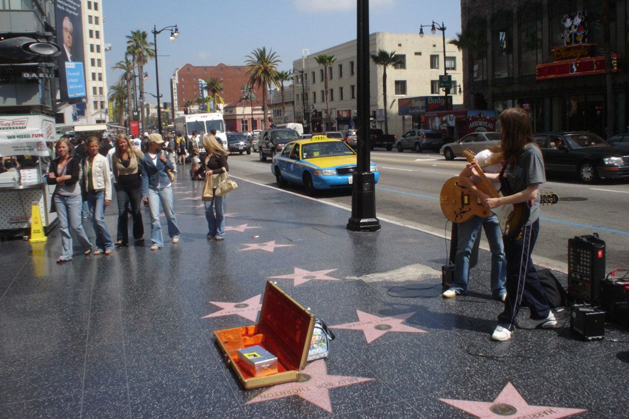 A street musician with long hair plays guitar near an open guitar case on the Hollywood Walk of Fame, as a group of people stand nearby and cars drive past palm trees and buildings in the background.