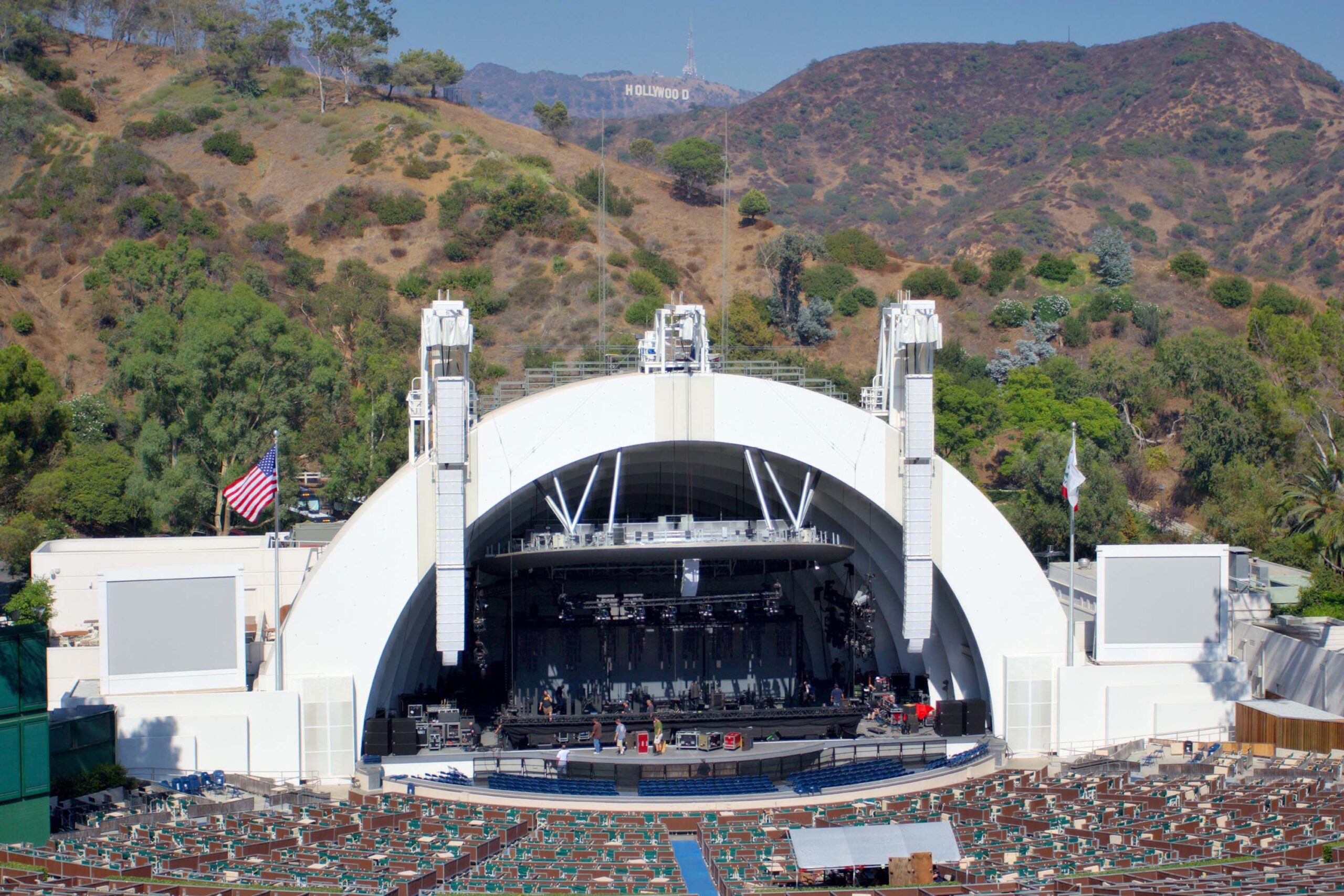 Outdoor amphitheater with white dome-shaped stage, rows of empty seats, American flag, and Hollywood hills with the Hollywood sign visible in the background.