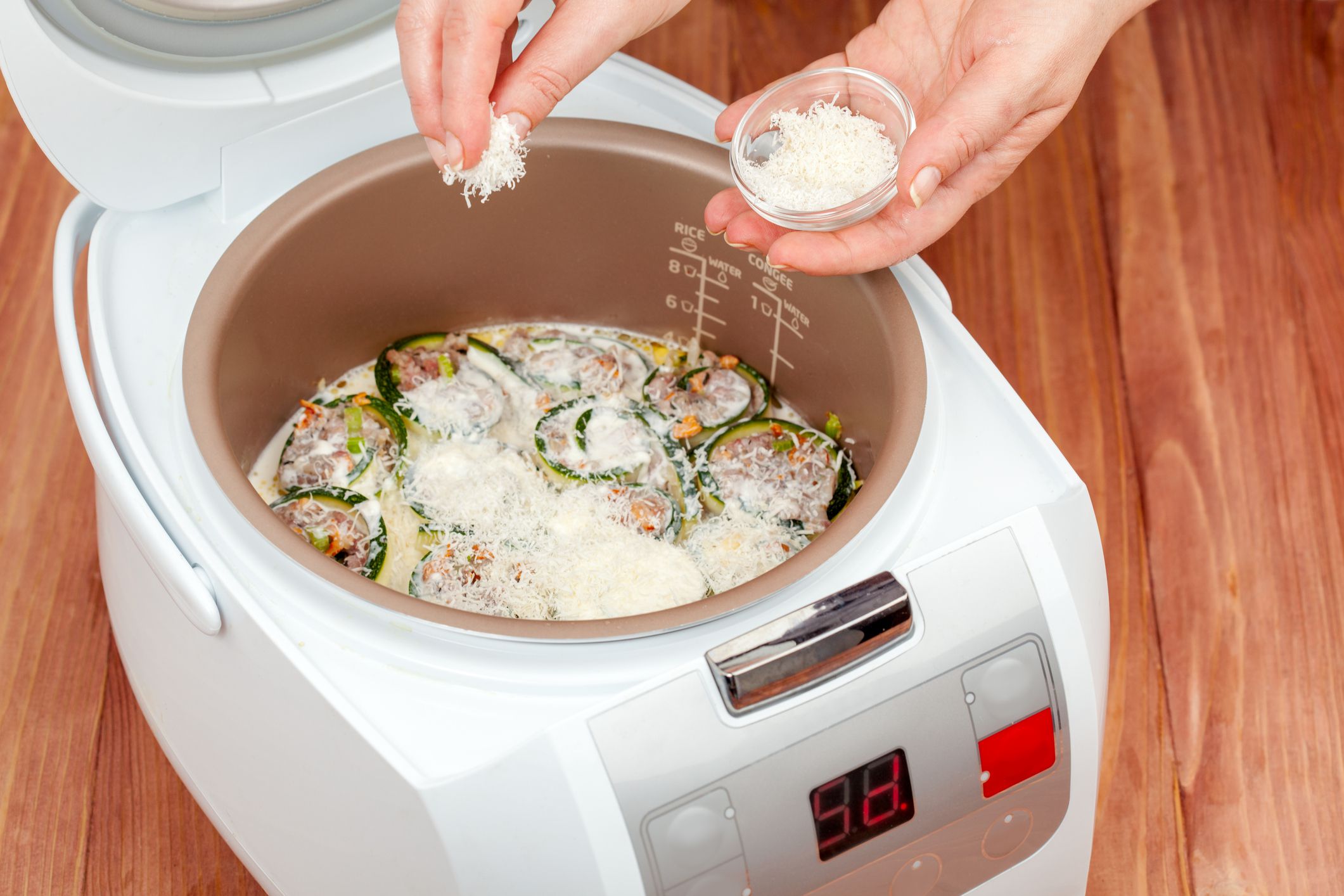 A person sprinkles shredded cheese from a small bowl into a rice cooker containing stuffed zucchini rounds in a creamy sauce, set on a wooden surface.