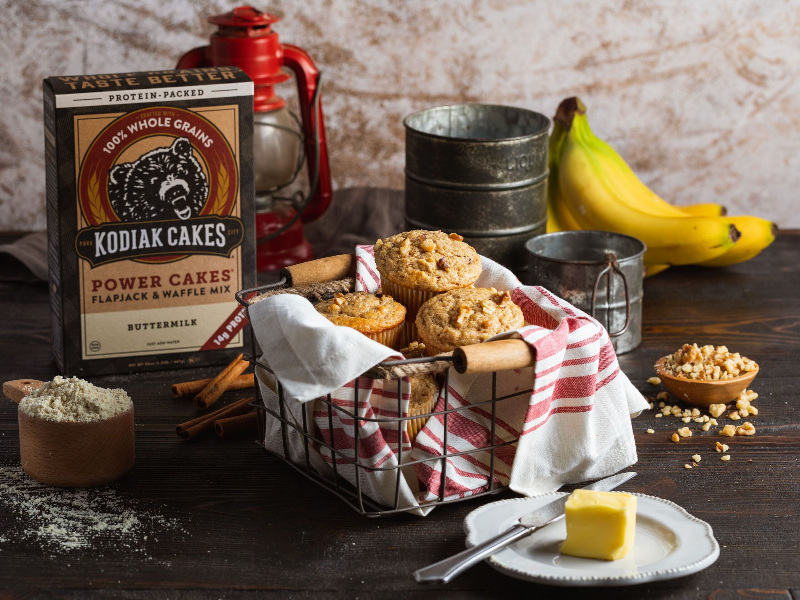 Kodiak Cakes Power Cakes Pancake and Waffle Mix with baked muffins in a metal basket, bananas, butter on a dark wood table