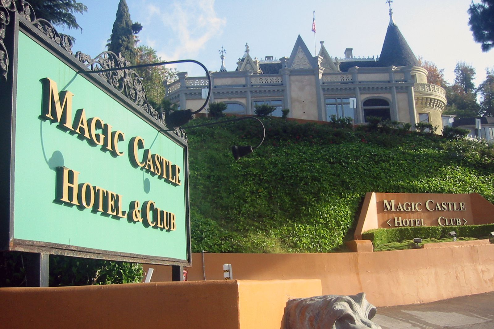 Large green and gold signs reading "Magic Castle Hotel & Club" in front of a historic, castle-like building on a hill covered with greenery, with a blue sky in the background.