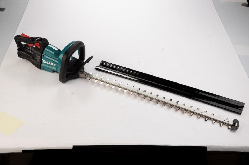 A Makita cordless hedge trimmer with a long silver blade and a black protective cover lying next to it, placed on a white surface.