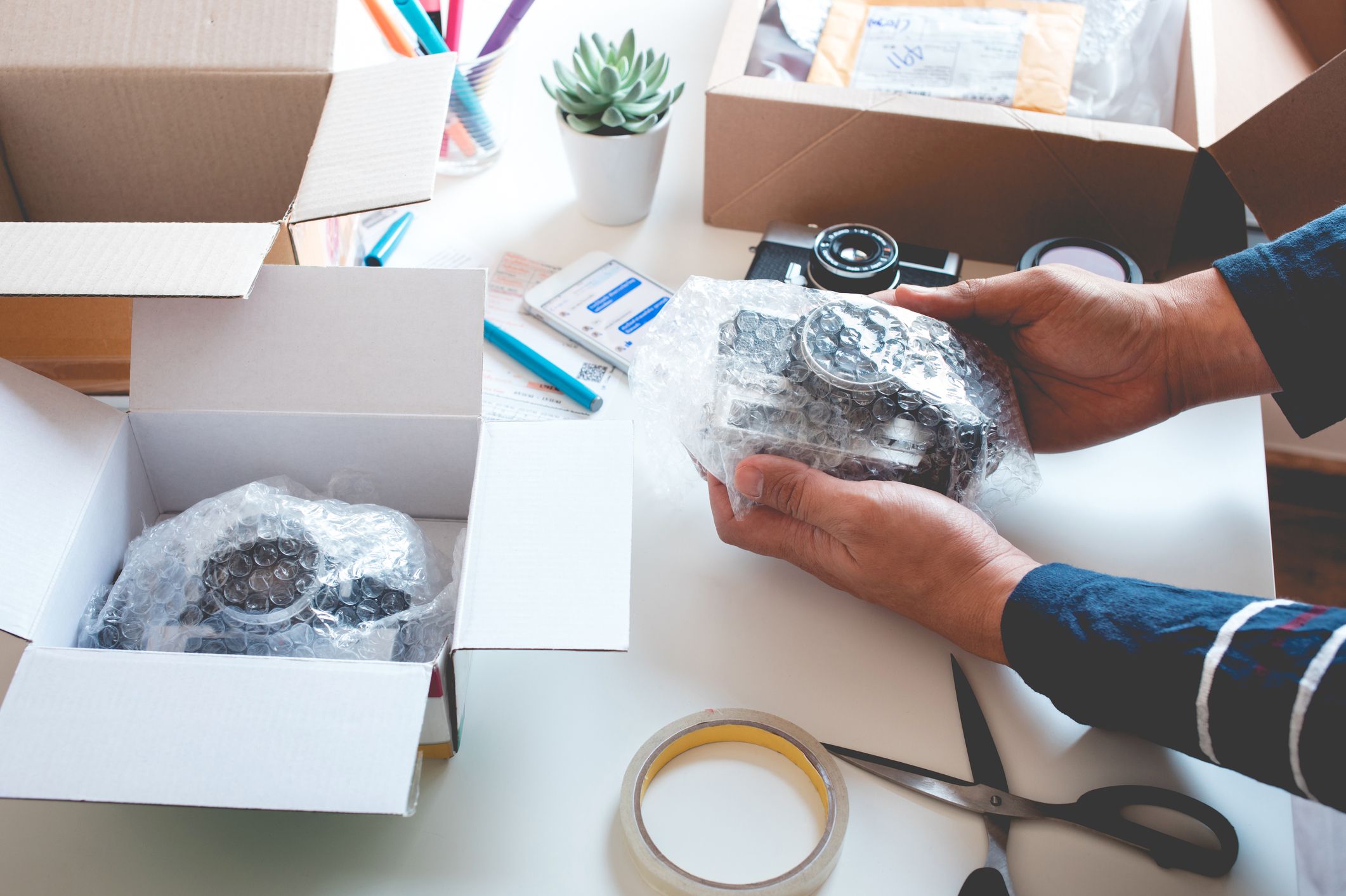 Male hands bubble wrapping camera for packaging to ship to customer, packaging materials on table, one shipping box is opened with another bubble wrapped camera for shipping