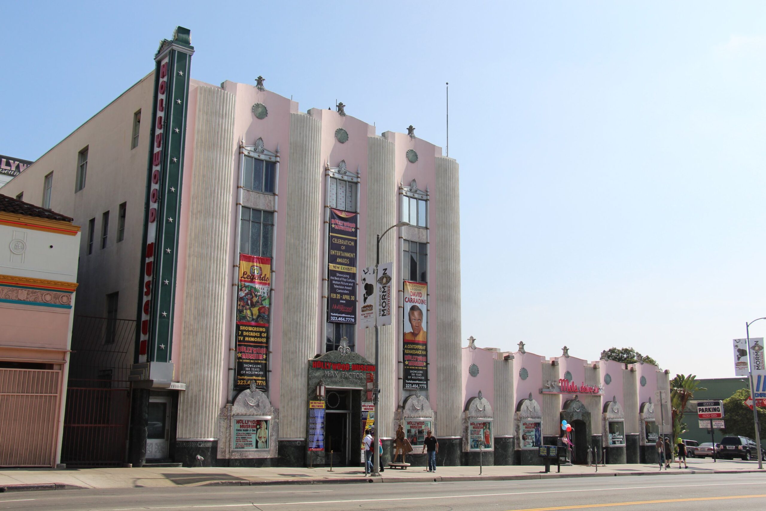 Art Deco style building with vertical lines, pink and white facade, tall central entrance, colorful event banners, and people walking on the sidewalk in front, under a clear sky.