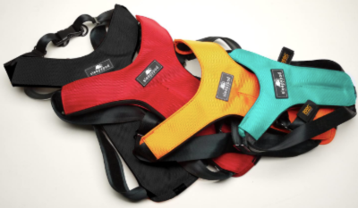 Three colorful dog harnesses—black, red, orange, and teal—are stacked on top of each other on a white background. Each harness has a black strap and a visible brand label.