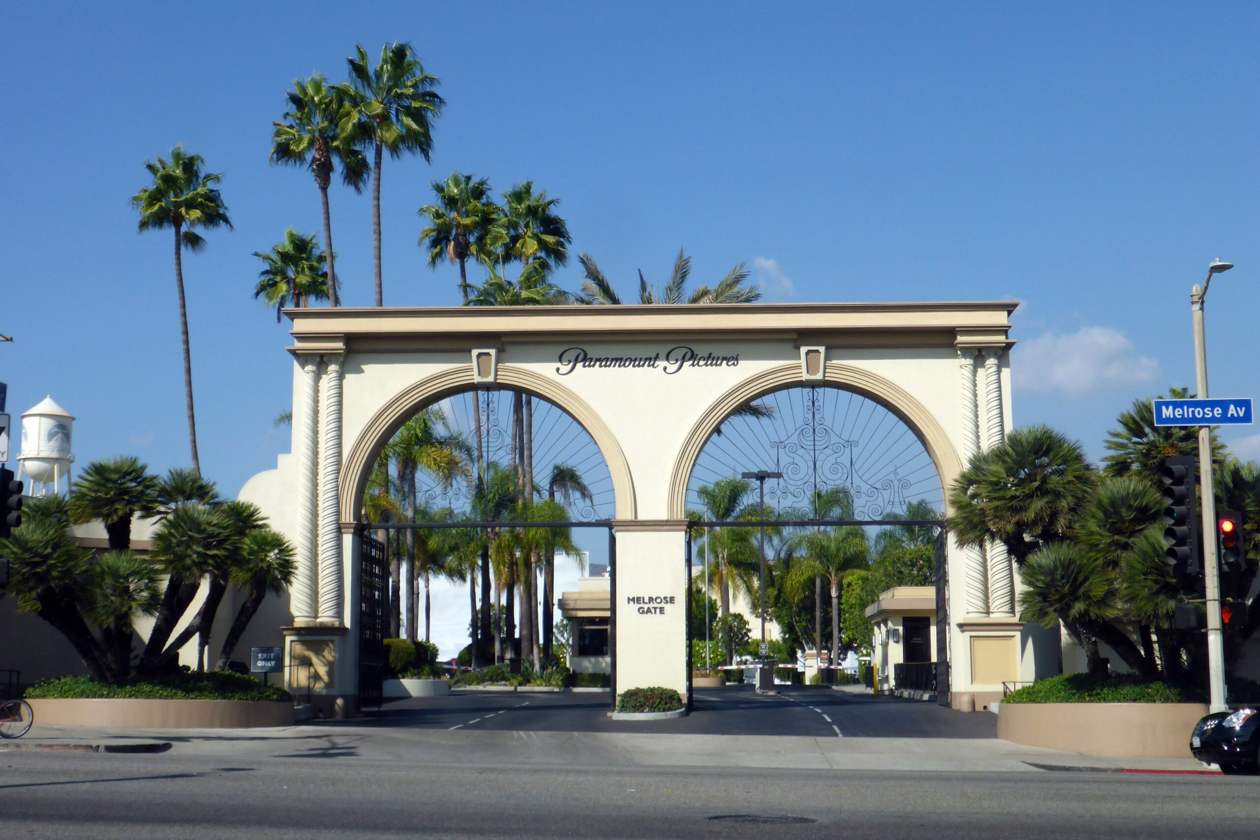 The iconic Paramount Pictures Melrose Gate, featuring ornate ironwork arches, palm trees, and a street sign for Melrose Avenue, under a clear blue sky.