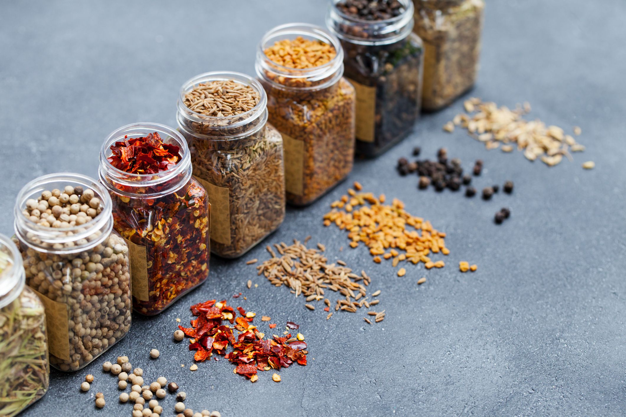 Assortments of spices, white pepper, chili flakes, lemongrass, coriander and cumin seeds in jars on grey stone background.