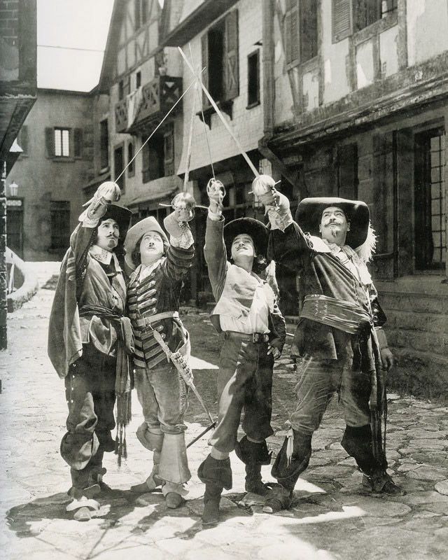 Four men in period costumes stand in a cobblestone street, raising their swords together in a salute. The scene appears to be from a historical or swashbuckling film set in old Europe.