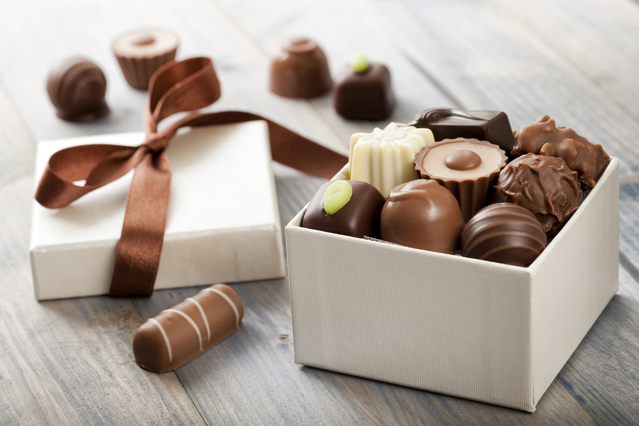 White box of fancy chocolates, mixed white, dark, and milk chocolates, box in the foreground, selective focus, on a grey wooden table, box top and five pieces of chocolates blurred in the background
