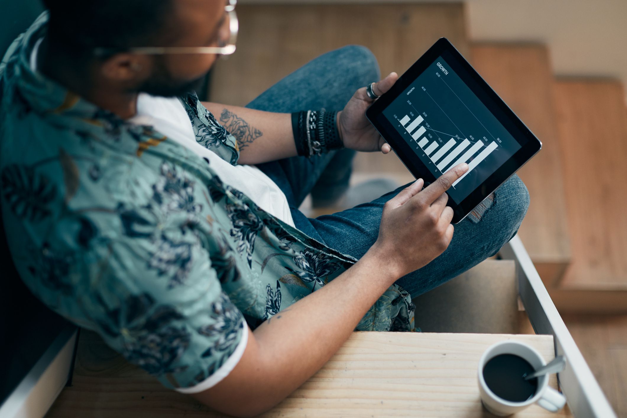 Young man on his stairs of his home, looking at his investments through a tablet, arm is resting on a stair and a cup of coffee on it