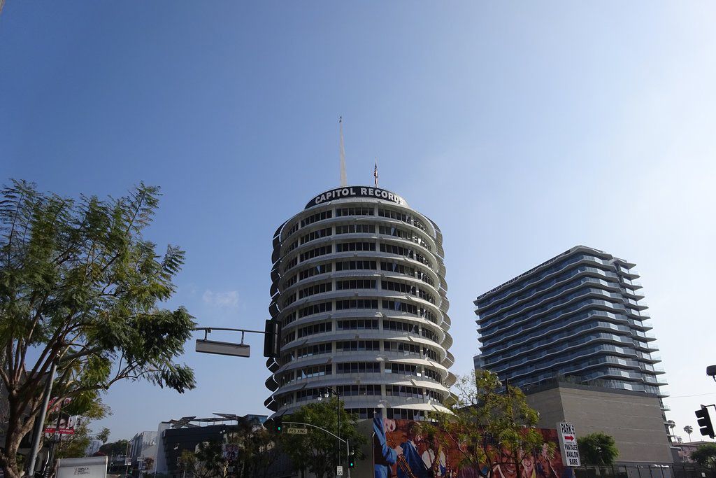 The image shows the iconic Capitol Records building in Los Angeles, a circular high-rise with a spire on top, against a clear blue sky. Nearby, there are trees and another modern building.