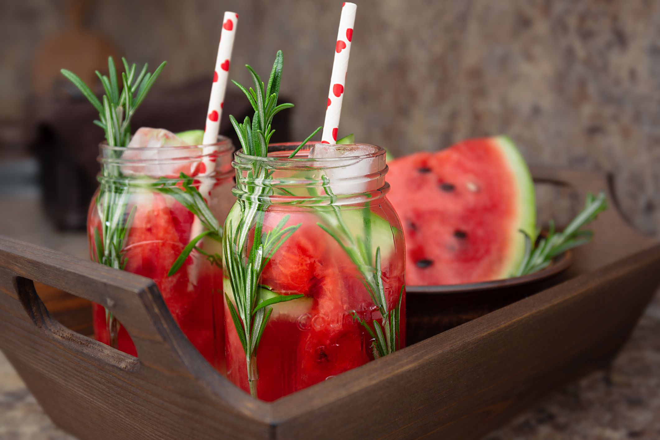 Two jars of refreshing summer drink with watermelon and rosemary served on a wooden tray. Watermelon and rosemary infused water with ice