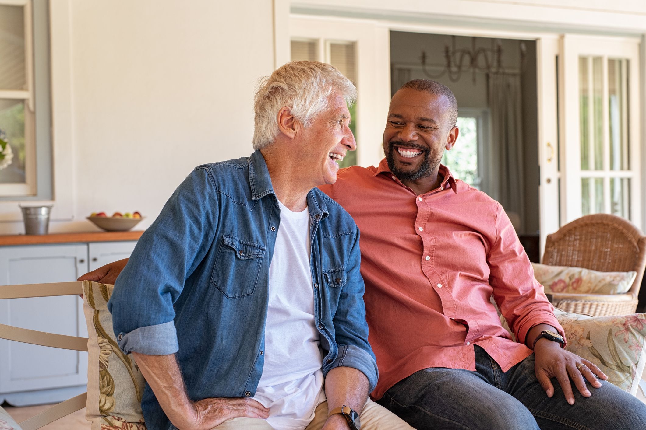 Two friends talking while sitting on couch in the courtyard. Senior man and african guy laughing while in conversation sitting outside home. Two men talking to each other and enjoying.