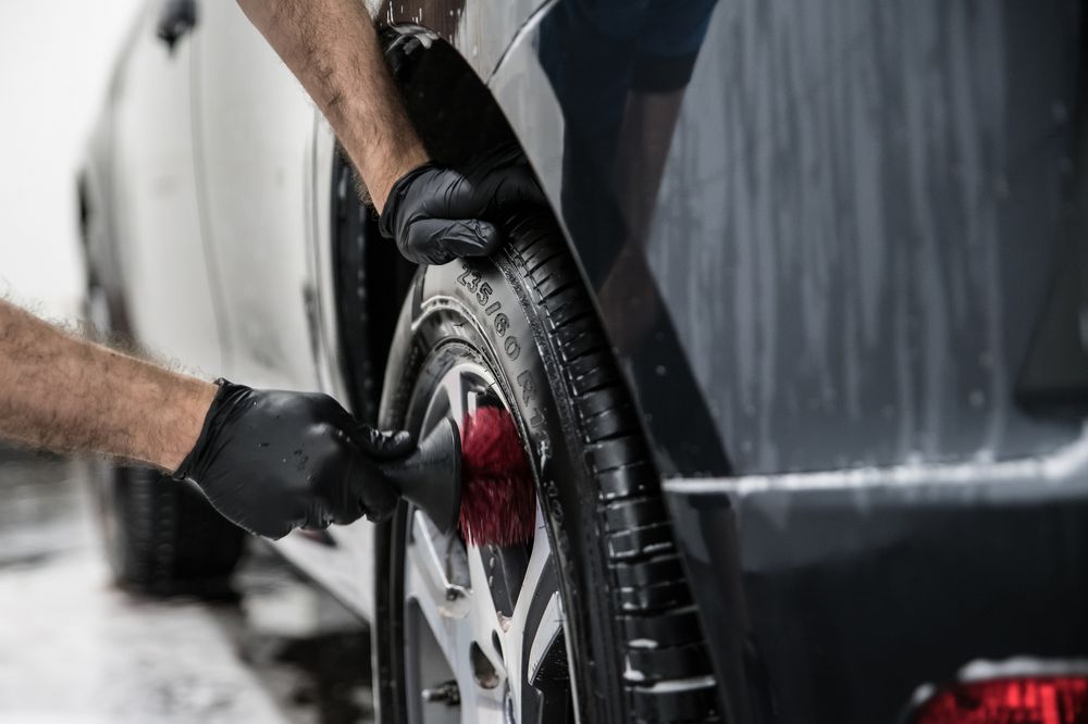 A person wearing black gloves uses a red brush to clean the tire and rim of a black car, which is wet and partially covered in soap.