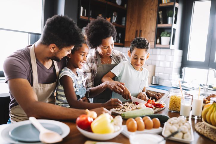 A family of four smiles and prepares food together in a kitchen, arranging toppings on a pizza with ingredients like vegetables and eggs visible on the counter.