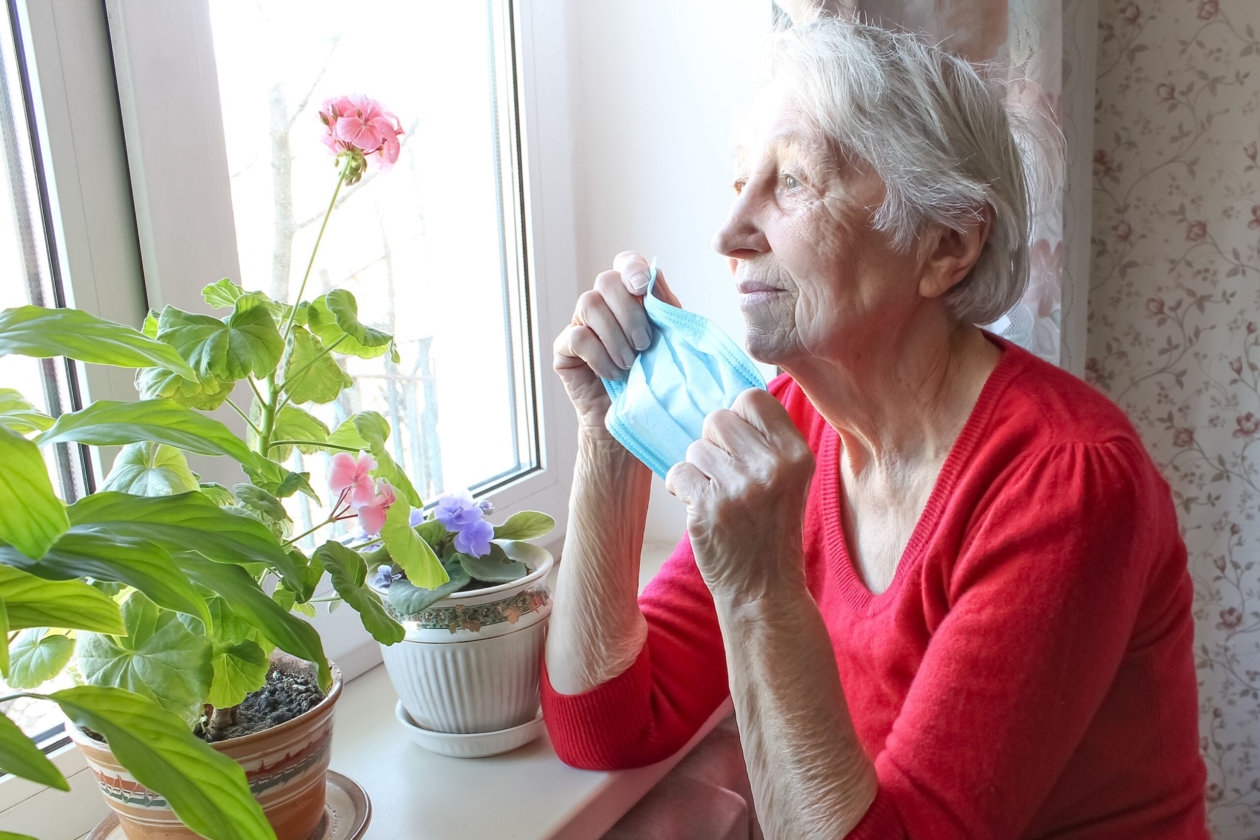 The Covid-19, health, safety and pandemic concept - senior old lonely woman wearing protective medical mask sitting near the window at home for protection from virus